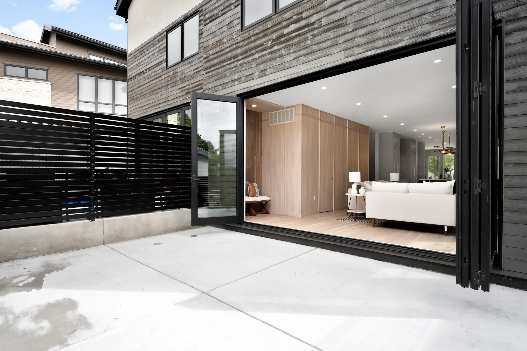 Open sliding glass door leading to a modern living room with light wood walls, white sofa, and black accents, outside concrete patio, black fence, and neighboring house