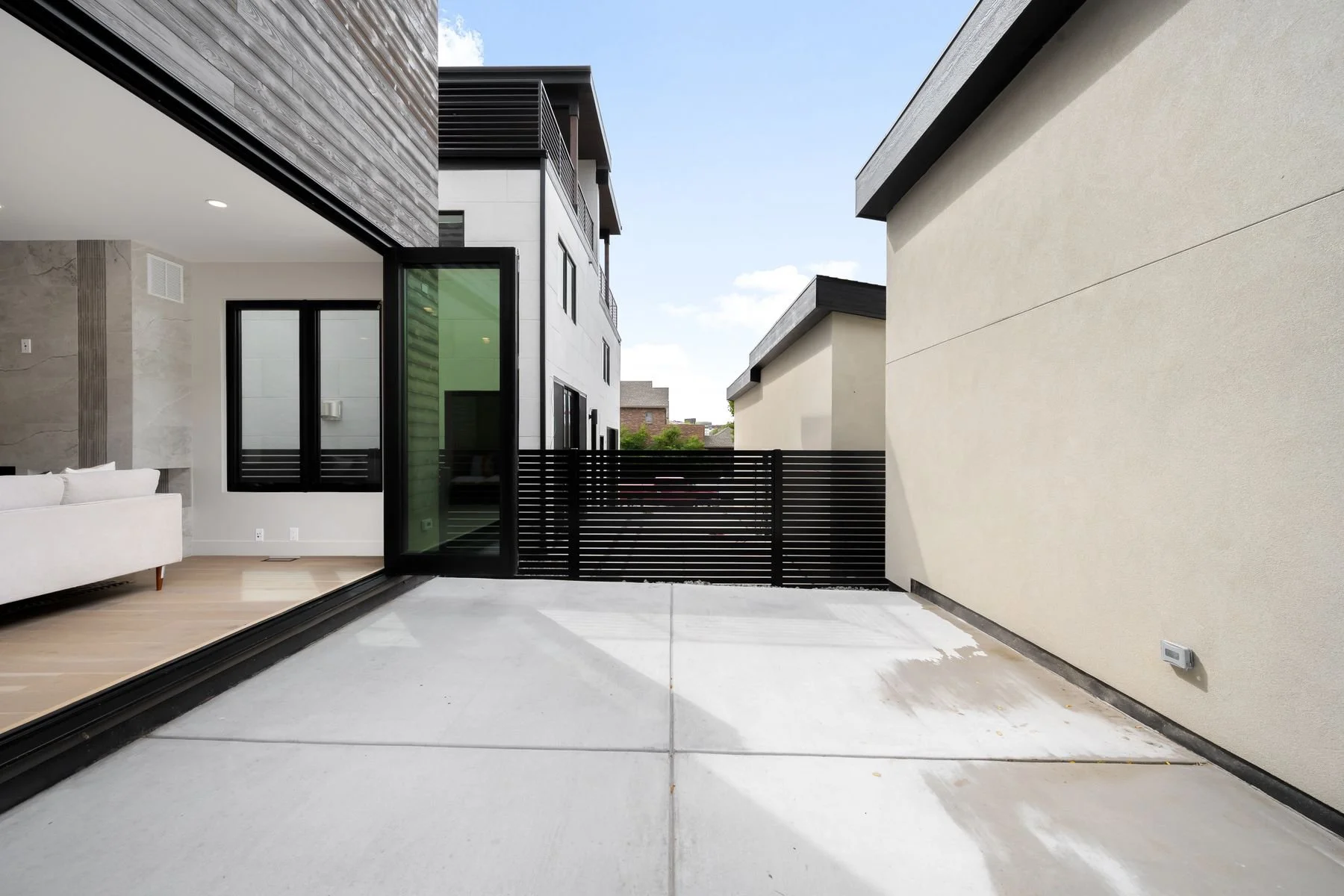 Front view of a modern house with a patio, black sliding door open to a living room, and neighboring buildings visible in the background.