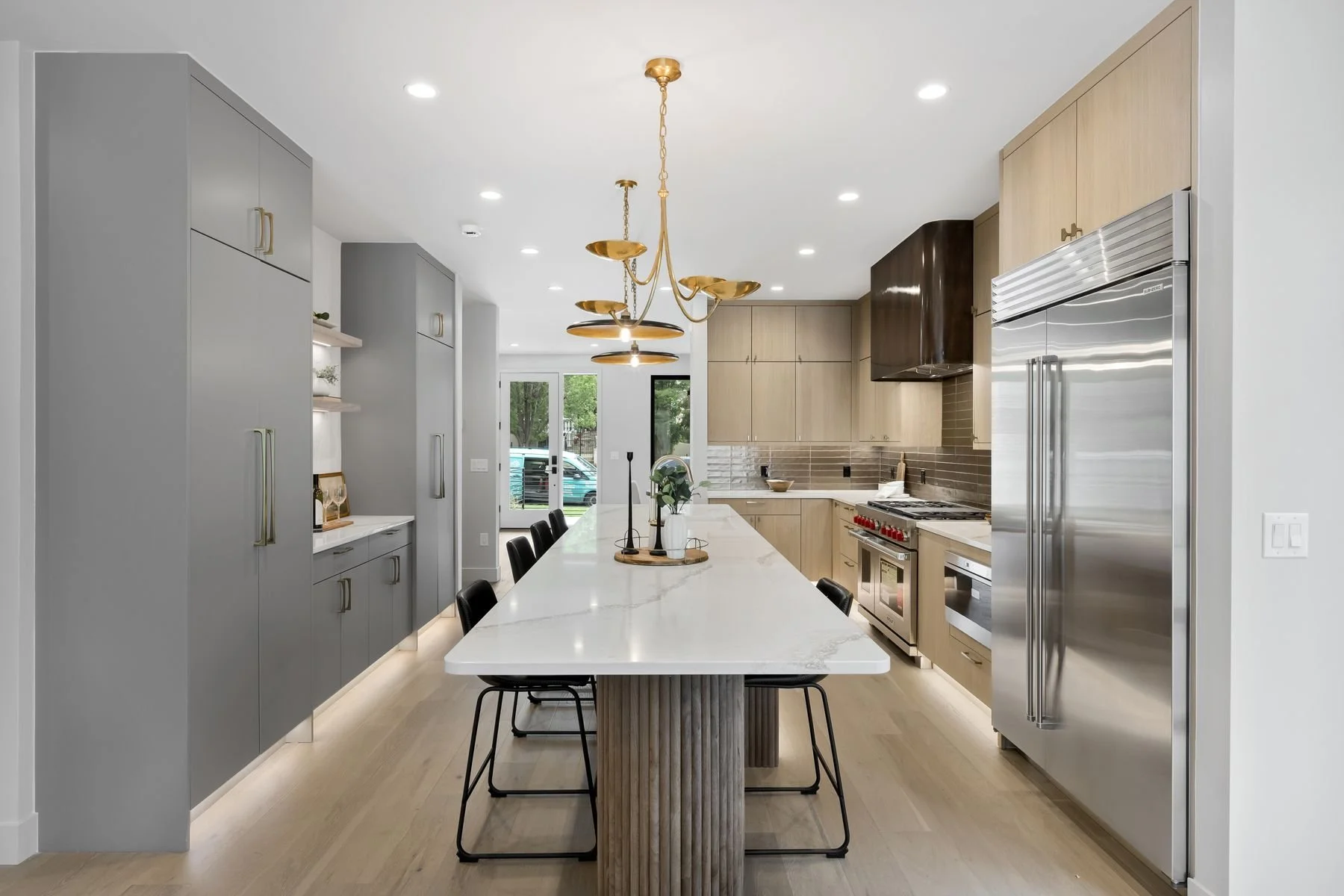 Modern kitchen with large white island, black chairs, gray cabinets, stainless steel appliances, wooden upper cabinets, beige tiled backsplash, and gold hanging light fixture.