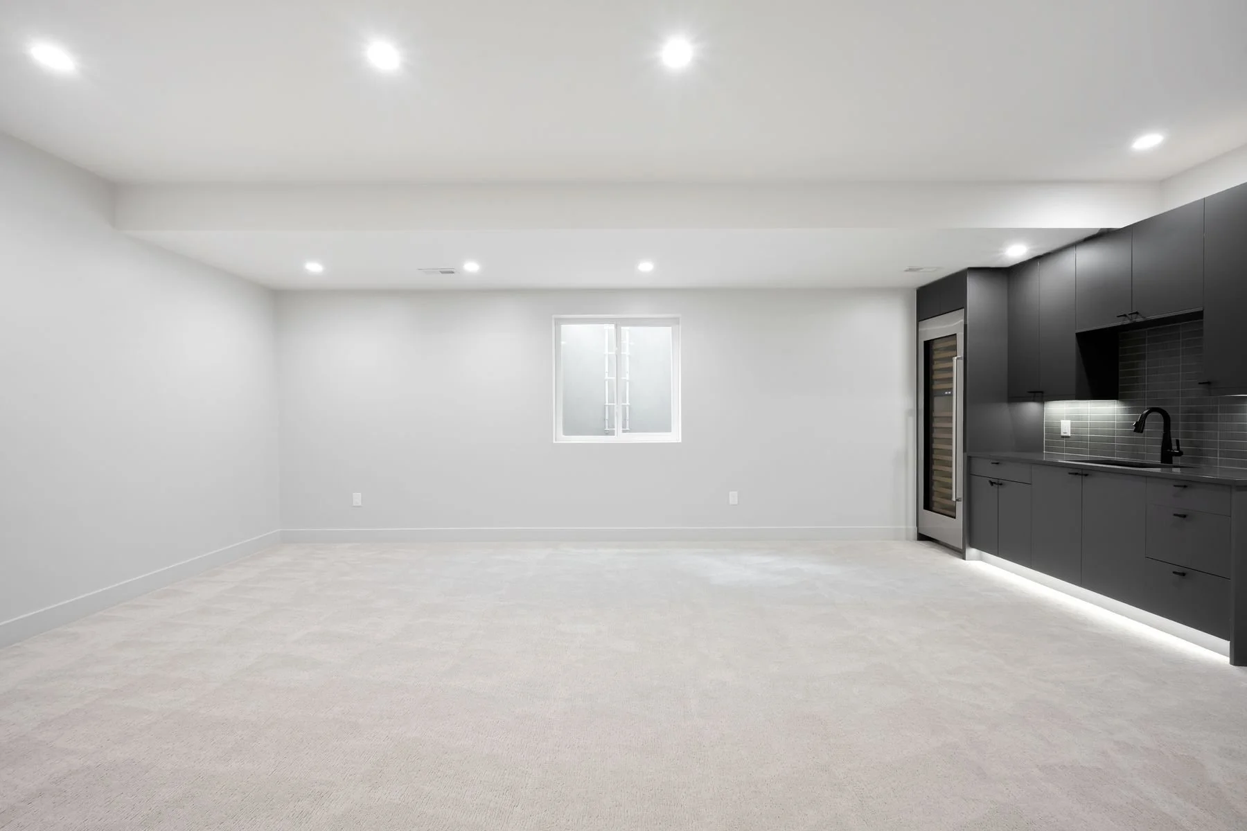 Empty modern basement with white walls, beige carpet, small window, and dark kitchen cabinets and appliances.