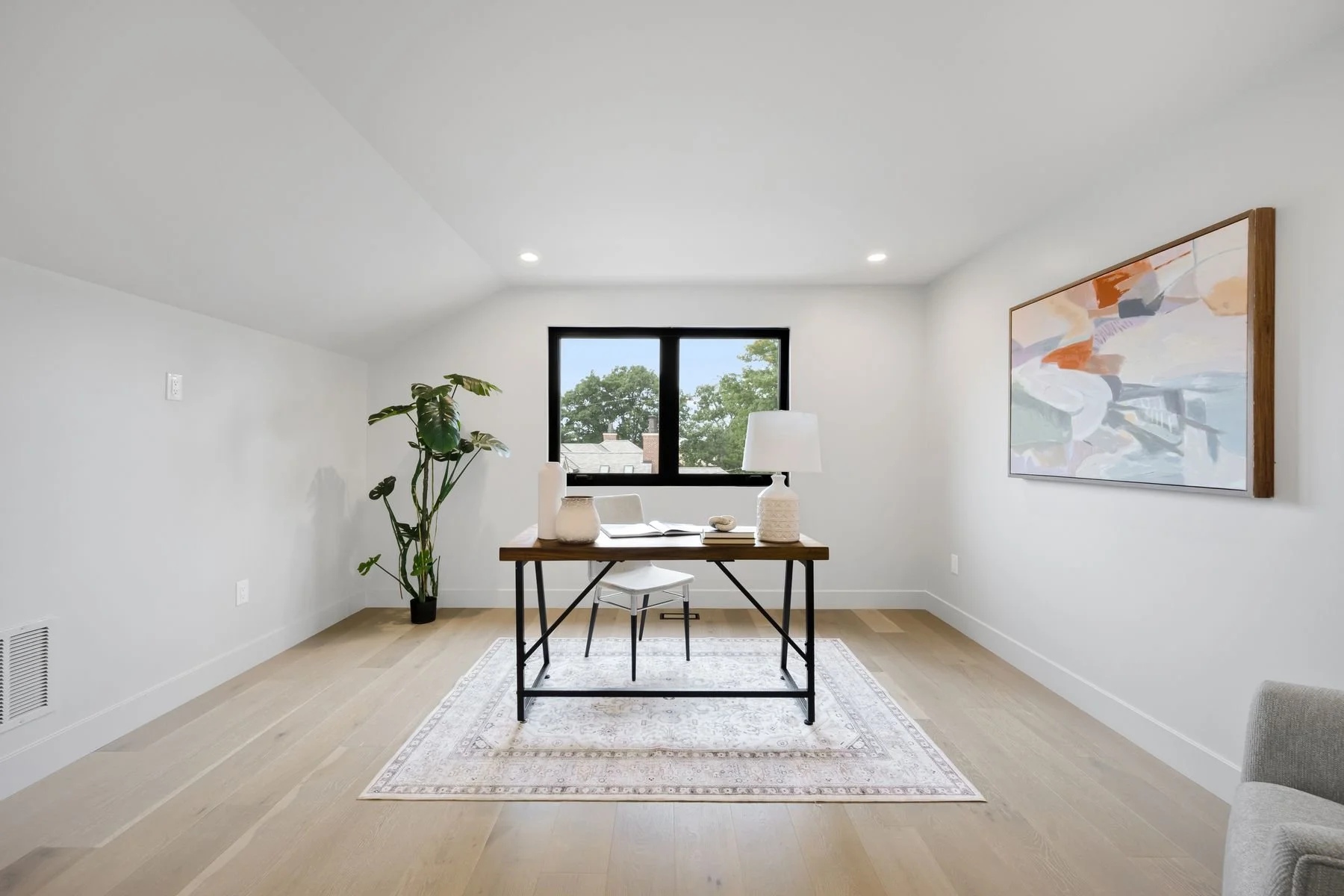 Minimalist home office with a wooden desk, white chair, black-framed window, potted plant, table lamp, open book, painting on the wall, and wooden flooring.