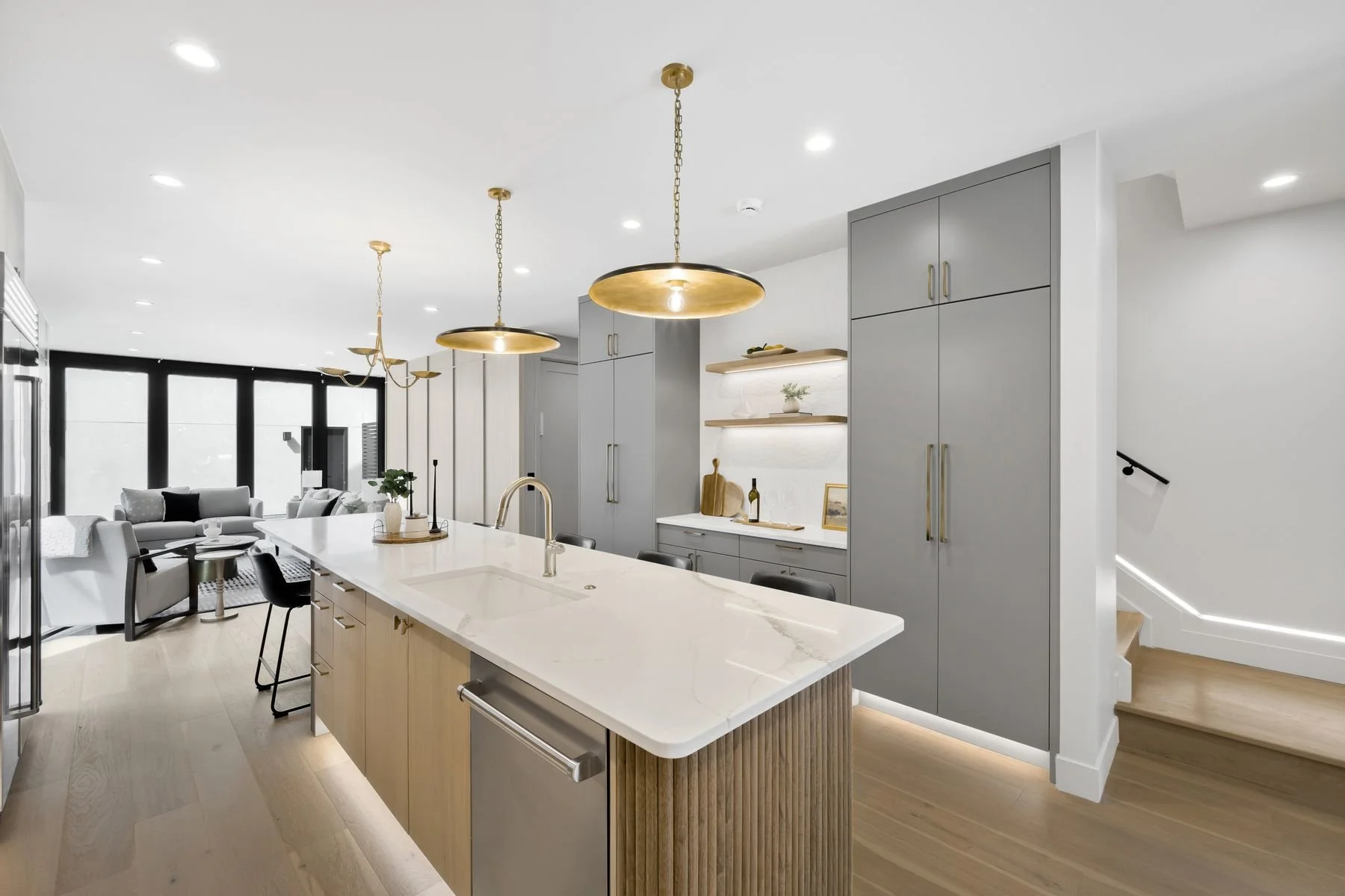 Modern open-concept kitchen with gray cabinetry, white marble island, and wood accents, leading to a living area with sofas and large windows.