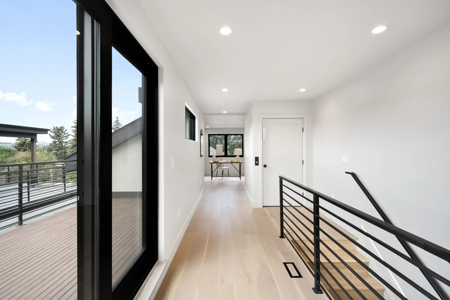 Modern interior hallway with white walls, wooden flooring, black metal railing stairs, and large sliding glass door leading to an outdoor balcony, with a small workspace visible at the end of the hall.