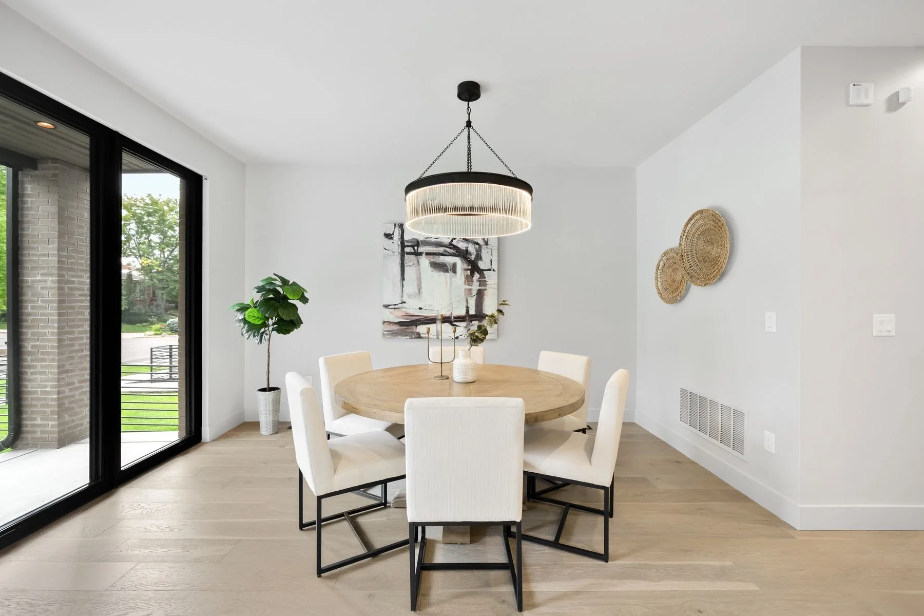 Modern dining room with a round wooden table, six white upholstered chairs, a black chandelier, abstract wall art, a potted plant, and natural light through large sliding glass doors.