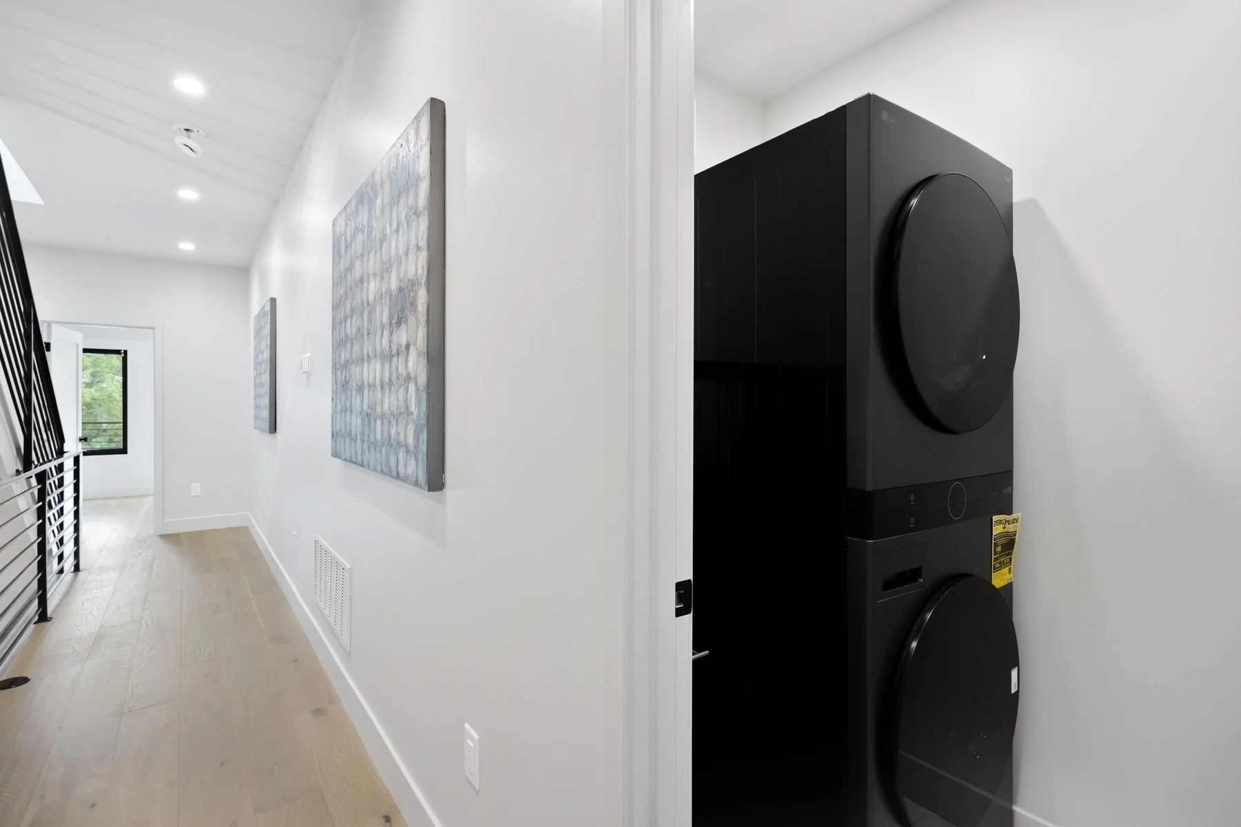A hallway with white walls and wooden floor leading to a room with a window; on the right side, there is a stacked black washer and dryer.