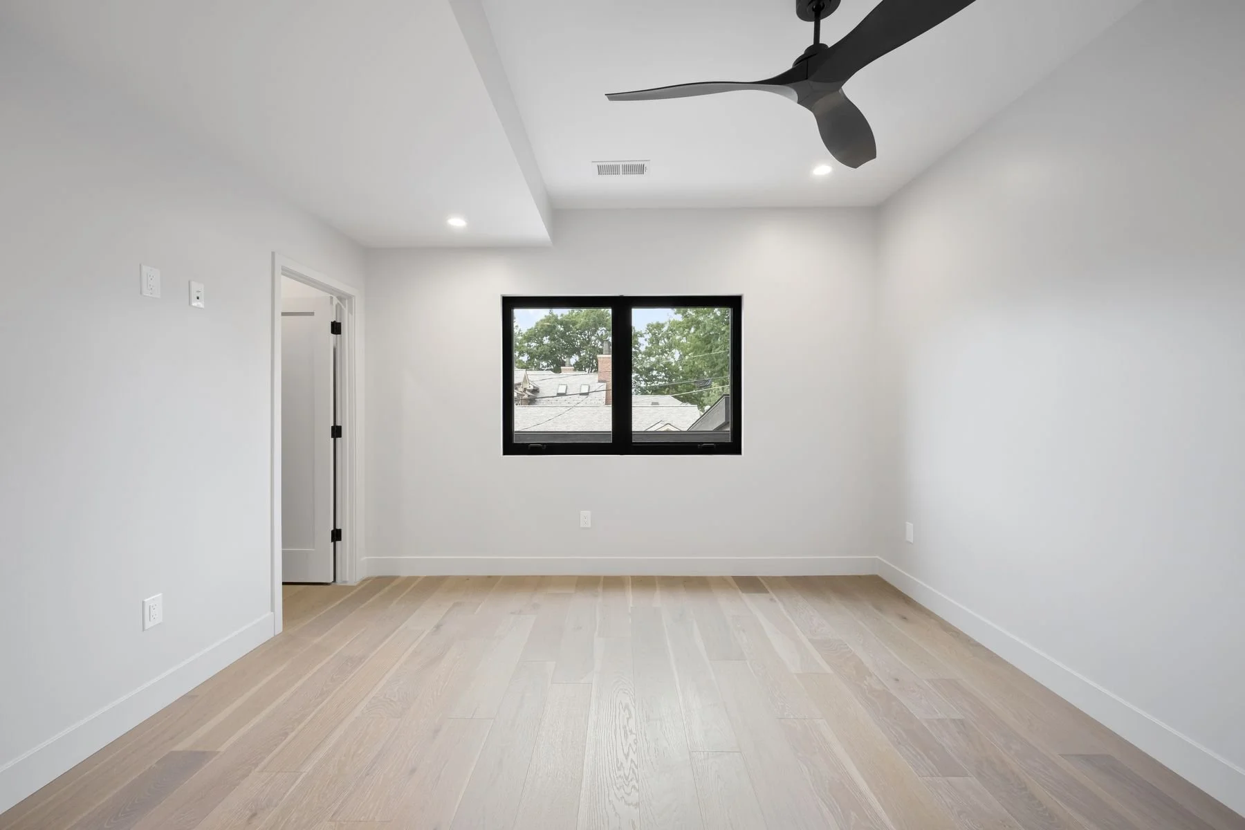 Empty room with white walls, light wooden floor, black window, ceiling fan, and door