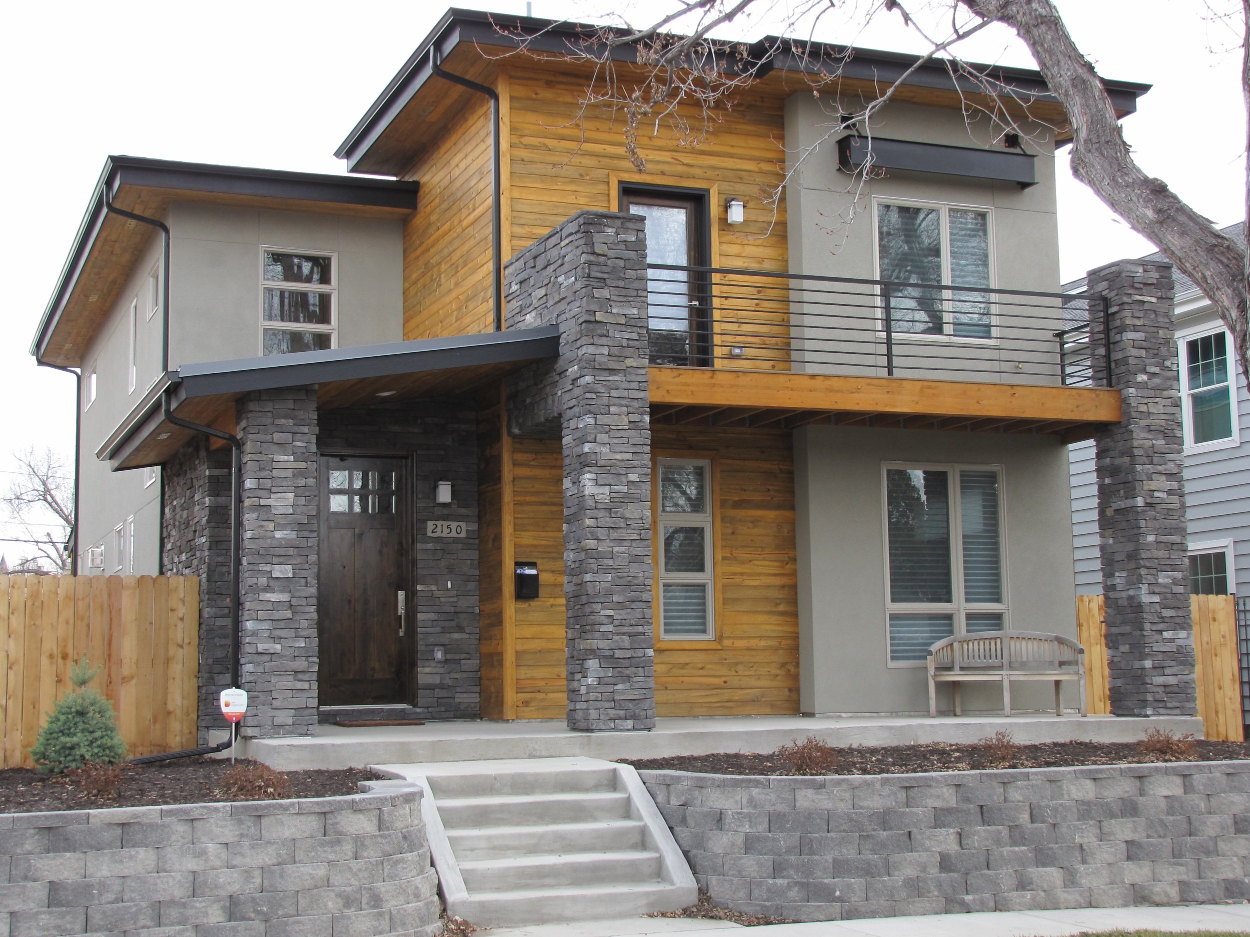 Modern two-story house with stone and wood exterior, front porch with stairs, and a bench.