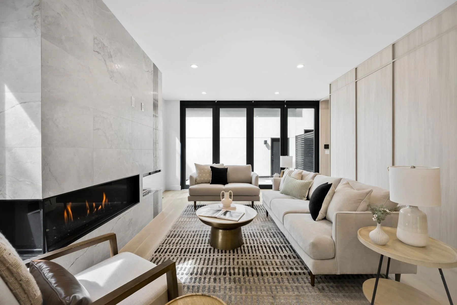 Modern living room with white sofas, a marble fireplace, large windowed doors, a round coffee table, and minimalist decor.