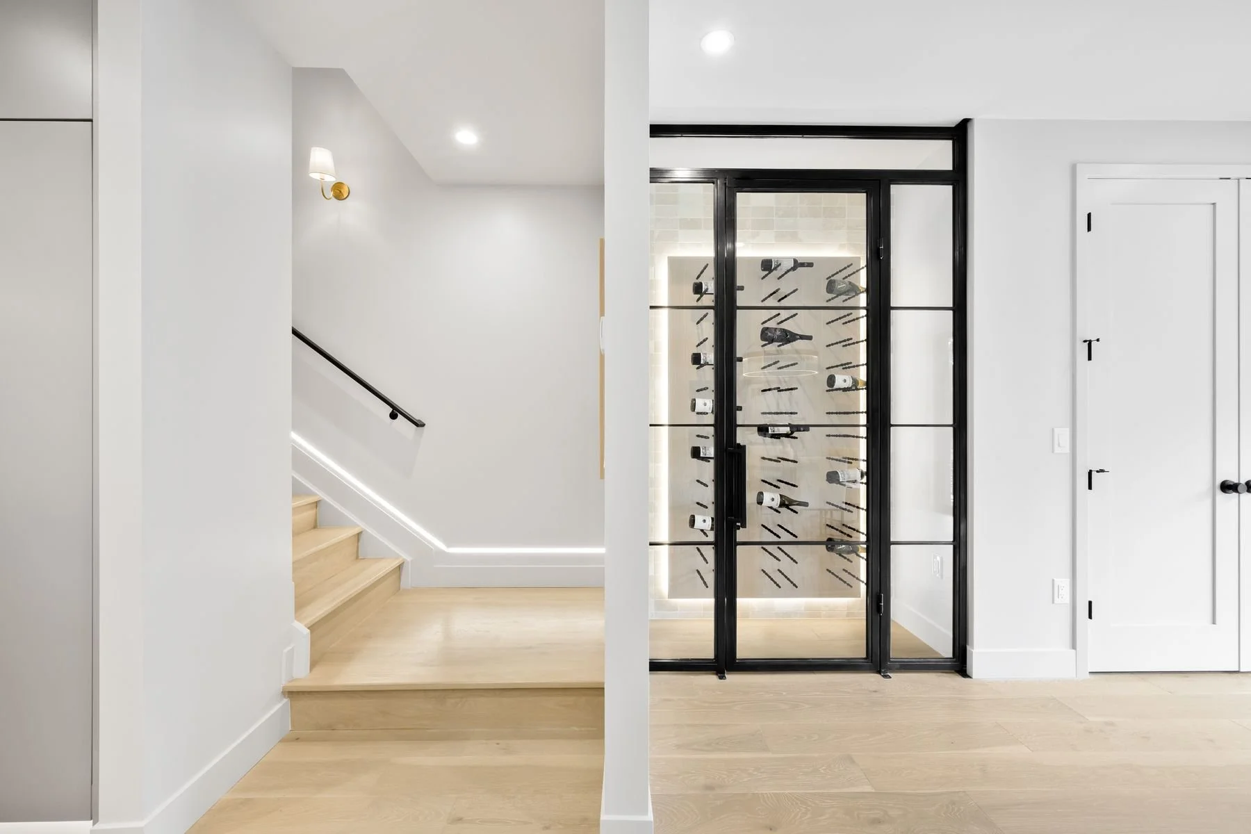 Interior of a modern home with a staircase on the left, white walls, and a wine cellar or collection display with a glass door on the right.