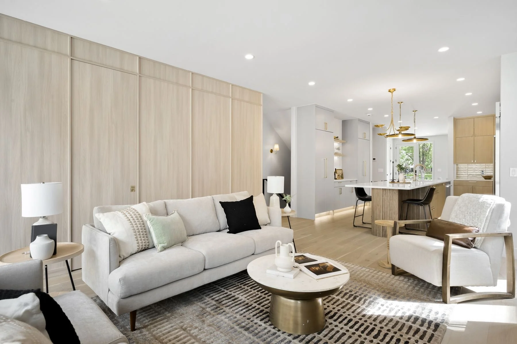 Modern living room with light-colored sofa, patterned rug, and a marble coffee table; open kitchen with island and gold lighting fixtures in the background.