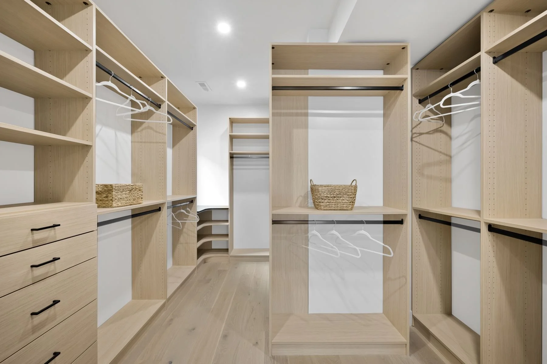 Empty walk-in closet with light wood shelves, drawers, and hanging rods, with a woven basket on a central shelf.
