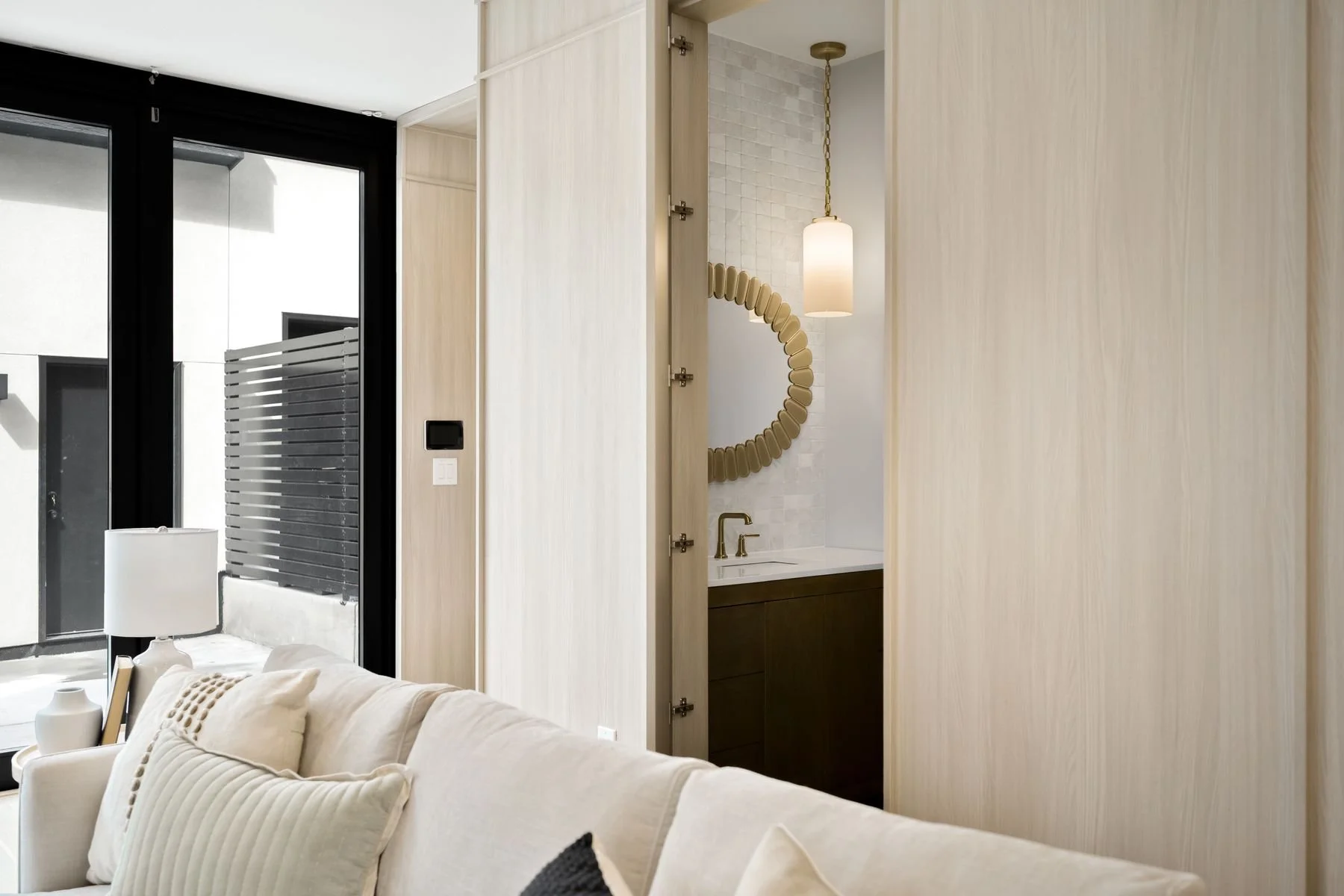 Interior of a modern living space with a beige couch, a side table with a lamp, and a sliding glass door leading outside. There is a small bathroom visible through an open door, featuring a round mirror, a hanging light, and a sink with a dark wooden