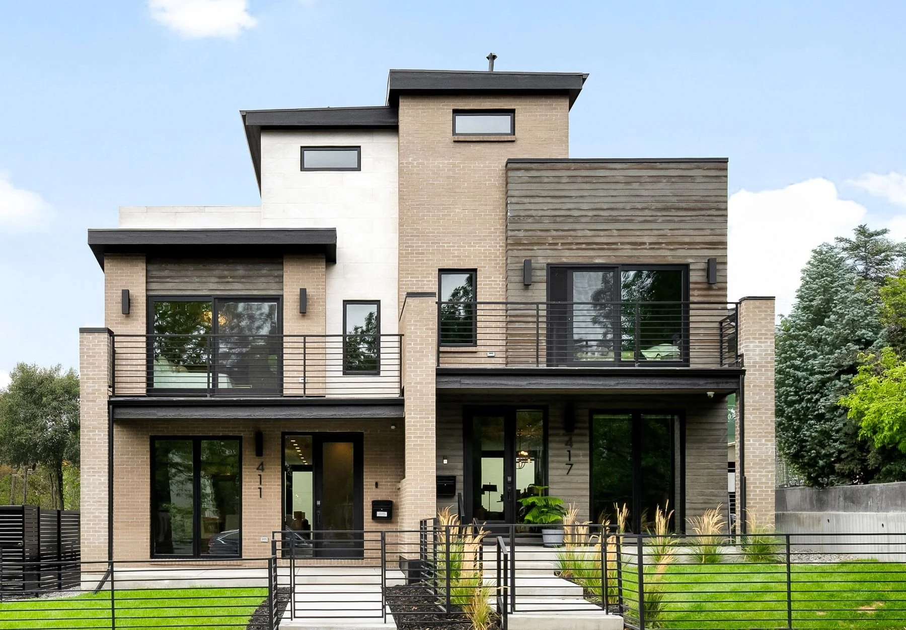 Modern multi-story house with brick and wood exterior, black railings on balconies, large windows, and a small front yard with green grass and bushes.