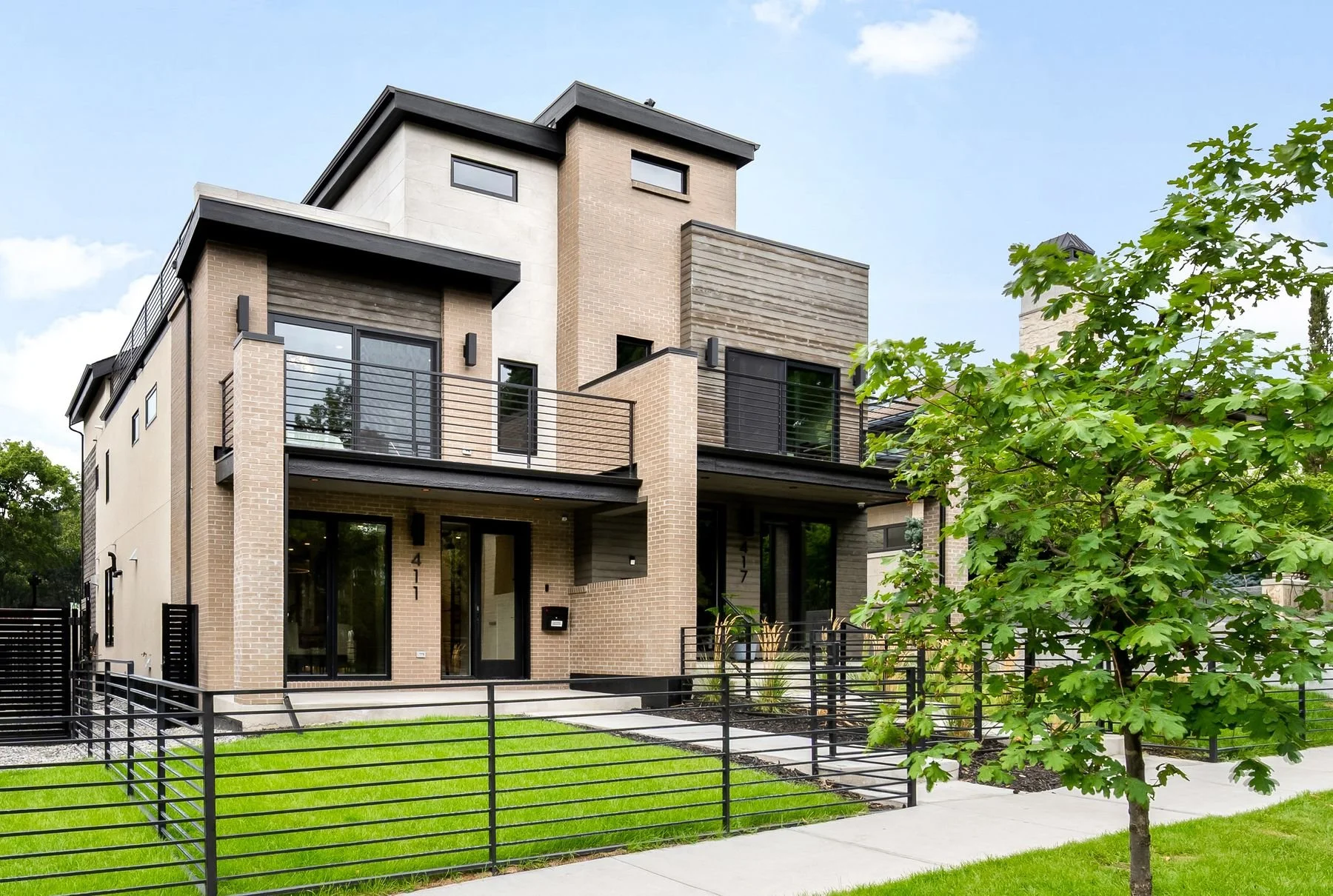 Modern multi-story house with brick and concrete exterior, black railings, and large windows, set on a green lawn with a sidewalk and small tree in the foreground.