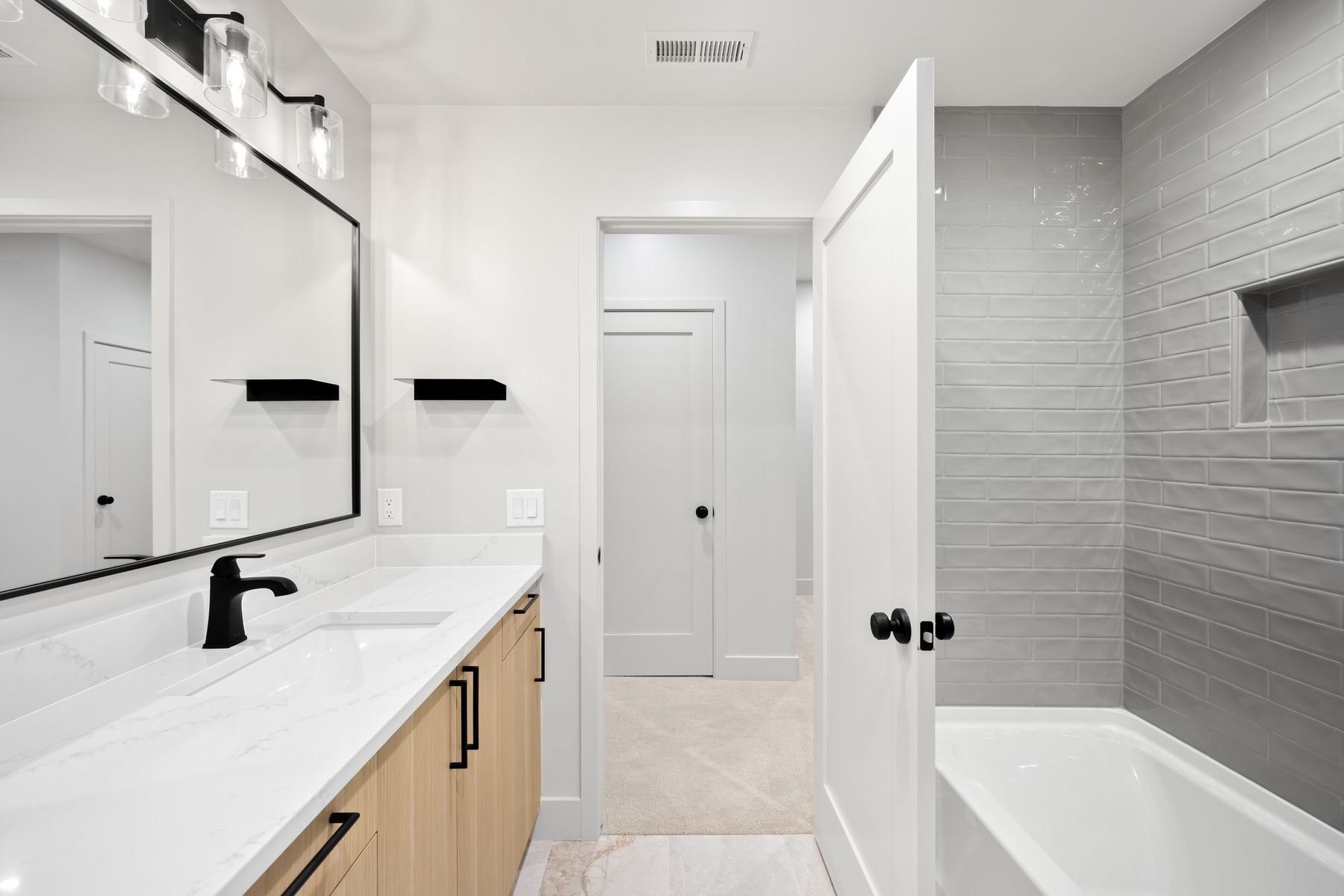 Modern bathroom with white walls, black fixtures, a large mirror, a wooden vanity with black handles, a white marble countertop, a black faucet, a gray tiled shower area with a niche, and a door leading to another room.
