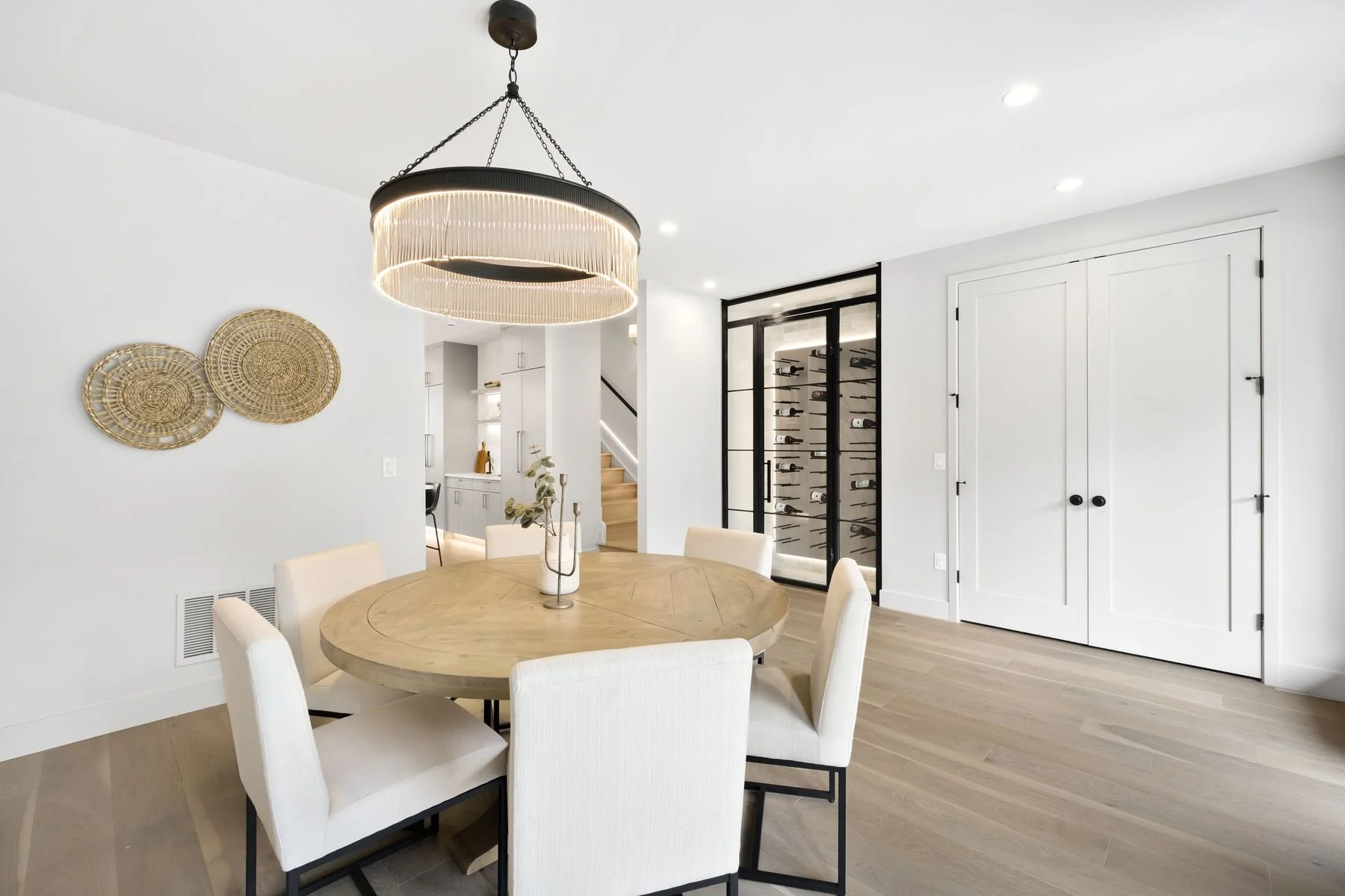 Modern dining room with a round wooden table, six white upholstered chairs, a black and gold chandelier, woven wall art, and a glass wine cellar with black framing in the background.