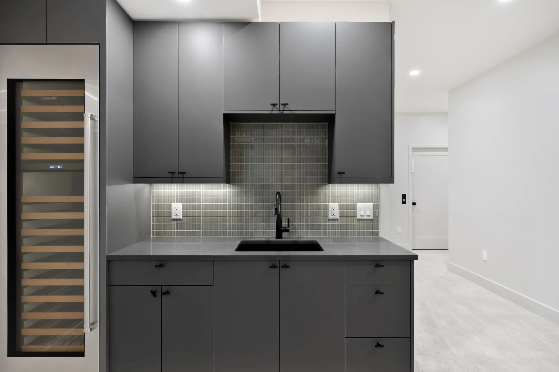 Modern kitchen with dark gray cabinets, a black sink, a gray countertop, a tiled backsplash, and a wine refrigerator on the left. There are electrical outlets and light switches on the backsplash area. The space is well-lit with recessed lighting, an