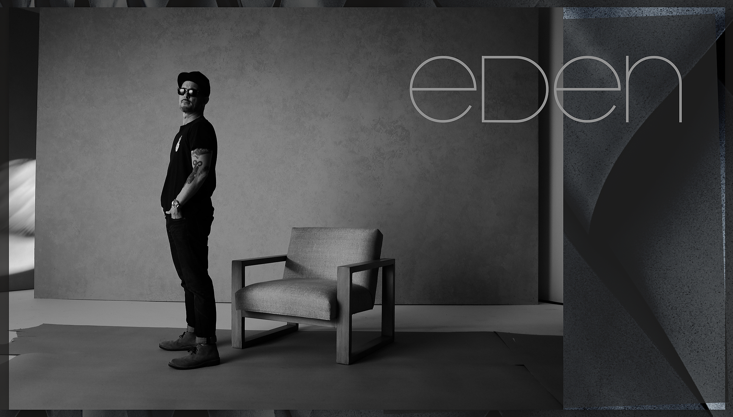 A man wearing sunglasses, a black t-shirt, black pants, and boots, standing beside a gray upholstered armchair in an art gallery or modern interior space with minimal decor and artwork on the wall.