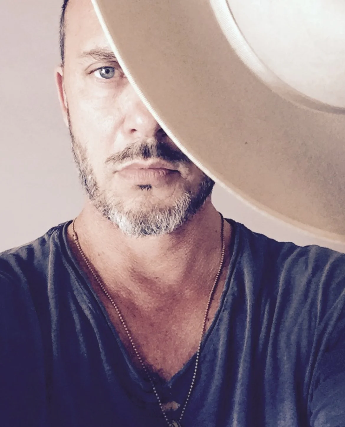 A man with a short beard and blue eyes, wearing a dark t-shirt and a necklace, partially obscured by a large beige hat.