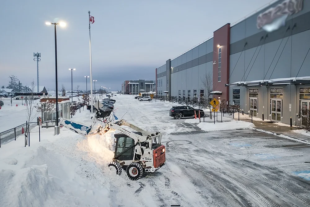 Skid-steer with a front mounted Top Gun Back Blade, moving snow in a parking lot.