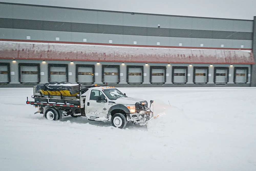 Snowcleared truck with a front plow and salter in the back, clearing an industrial loading bay.