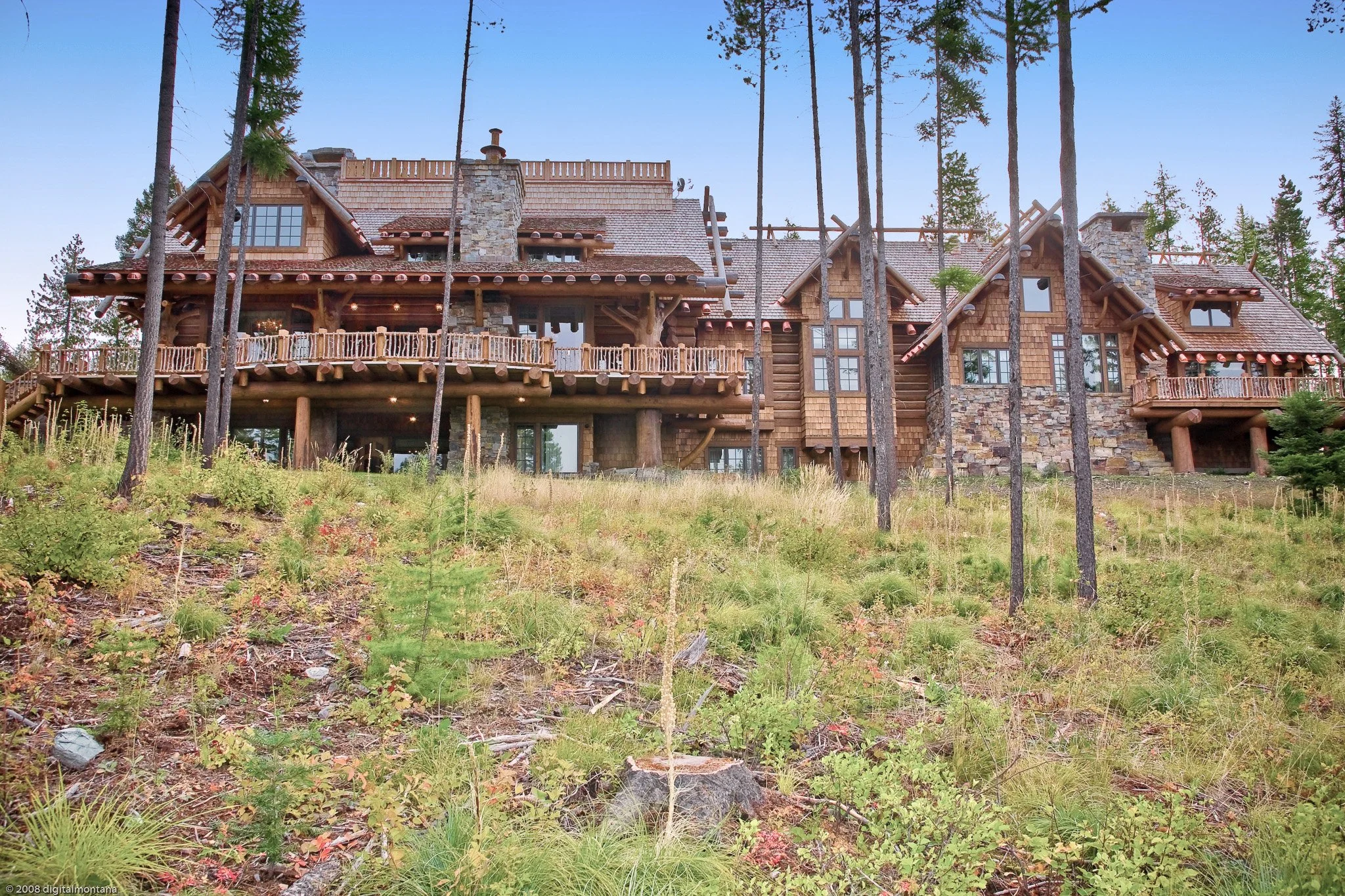 A large wooden house in the woods with tall trees surrounding it, multiple levels, a stone chimney, and a spacious balcony
