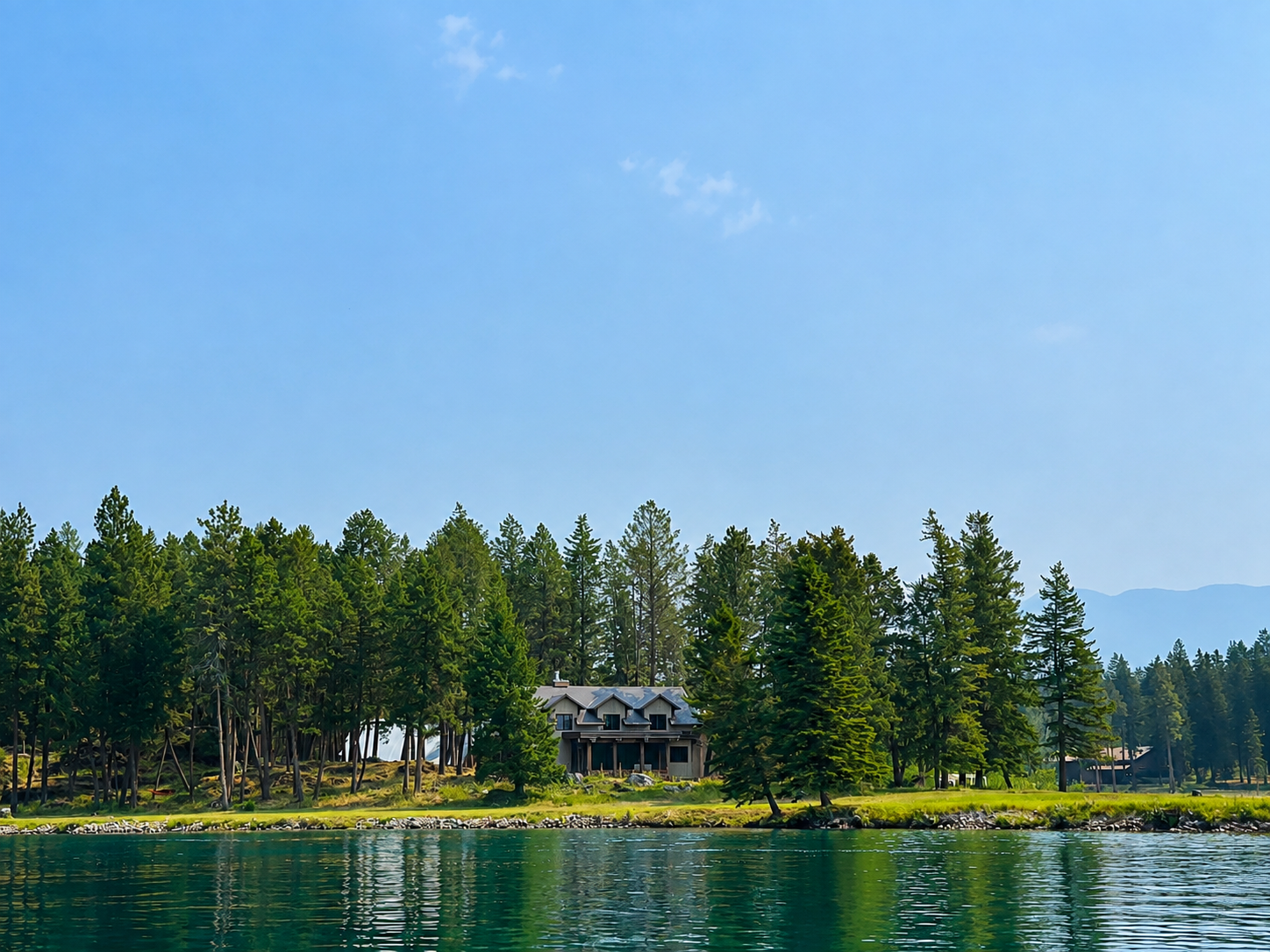 A house on a riverfront surrounded by pine trees, with mountains in the background and a clear blue sky overhead.