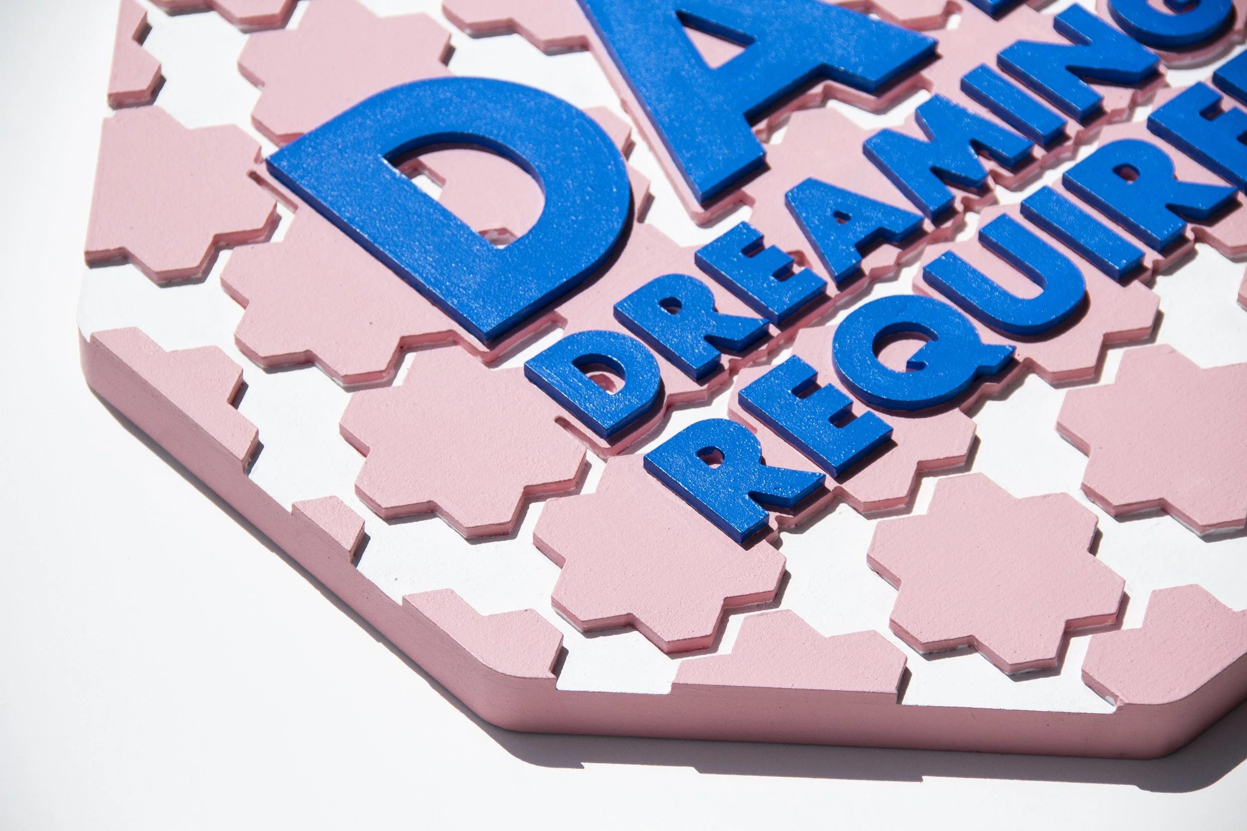 Detail An image of an imitation street sign that says "Day Dreaming Required" in blue with soft pink and white shapes.