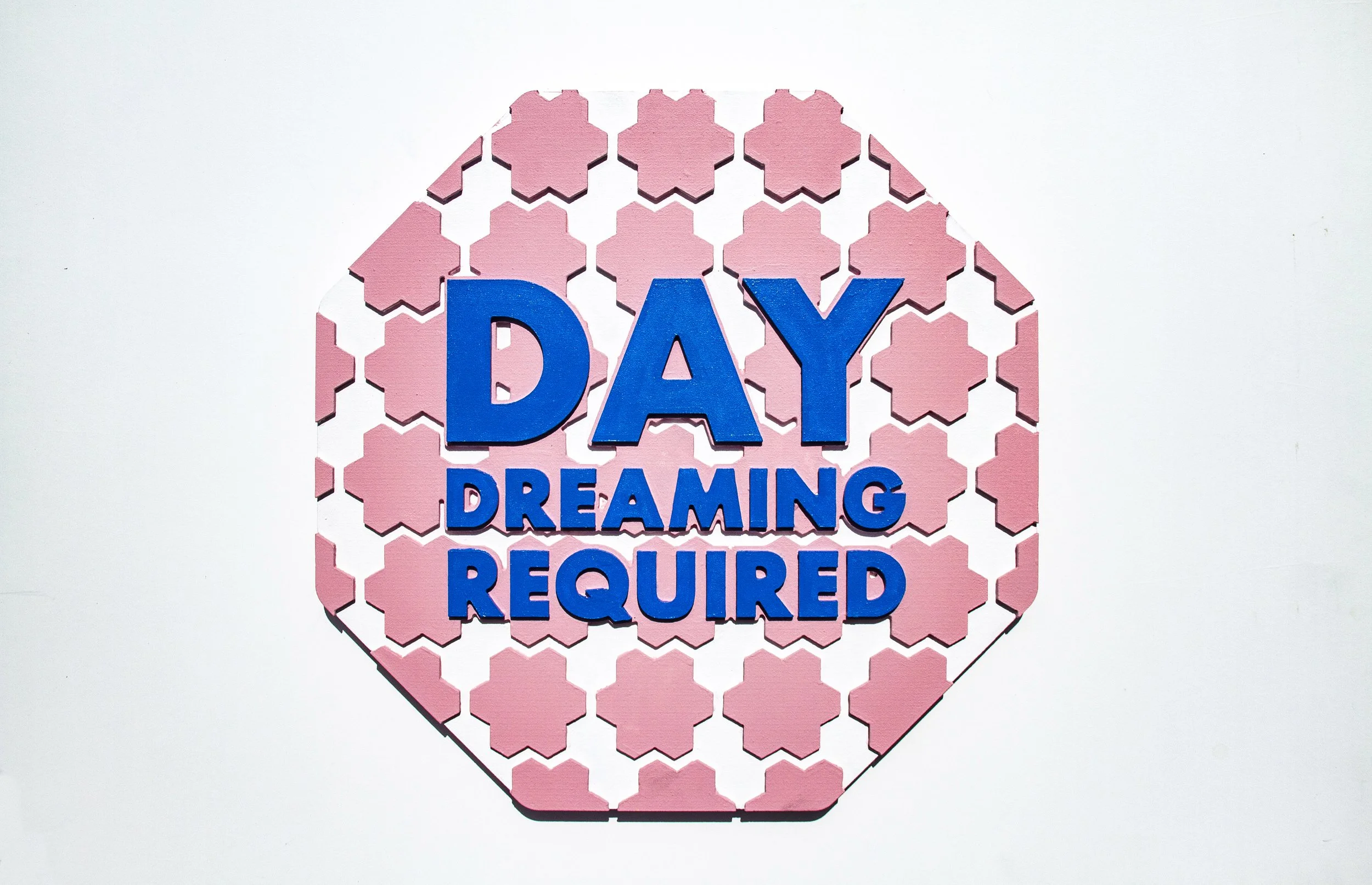 An image of an imitation street sign that says "Day Dreaming Required" in blue with text and soft pink and white shaped background.
