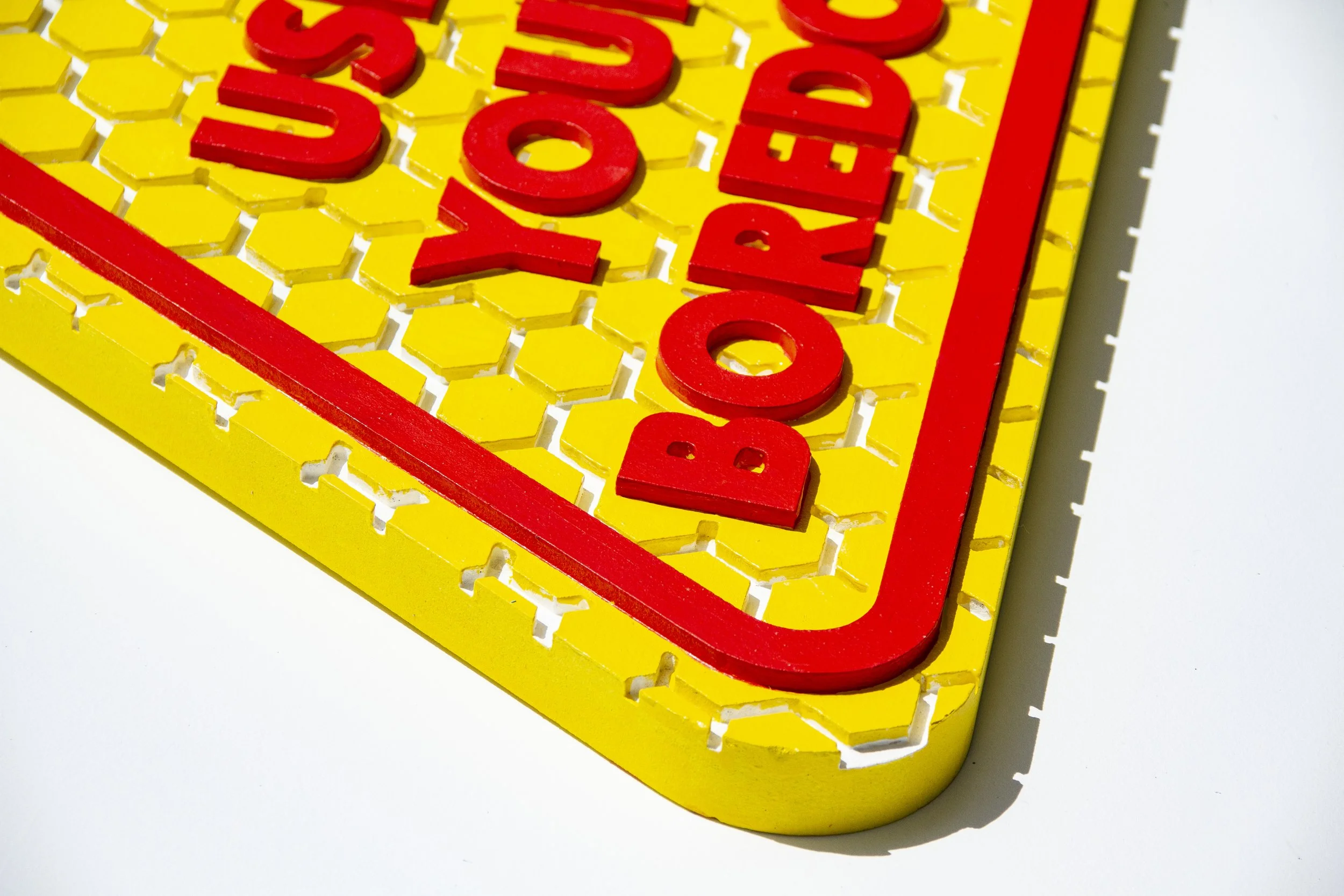 A detail image of an imitation street sign that says "Use Your Boredom"  with bright red text and outline line, with yellow hexagons with traces of white showing.