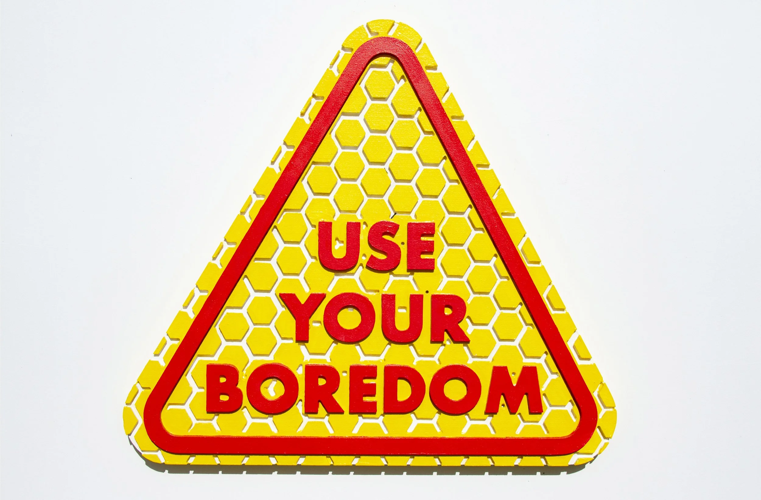 An image of an imitation street sign that says "Use Your Boredom"  with bright red text and outline line, with yellow hexagons with traces of white showing