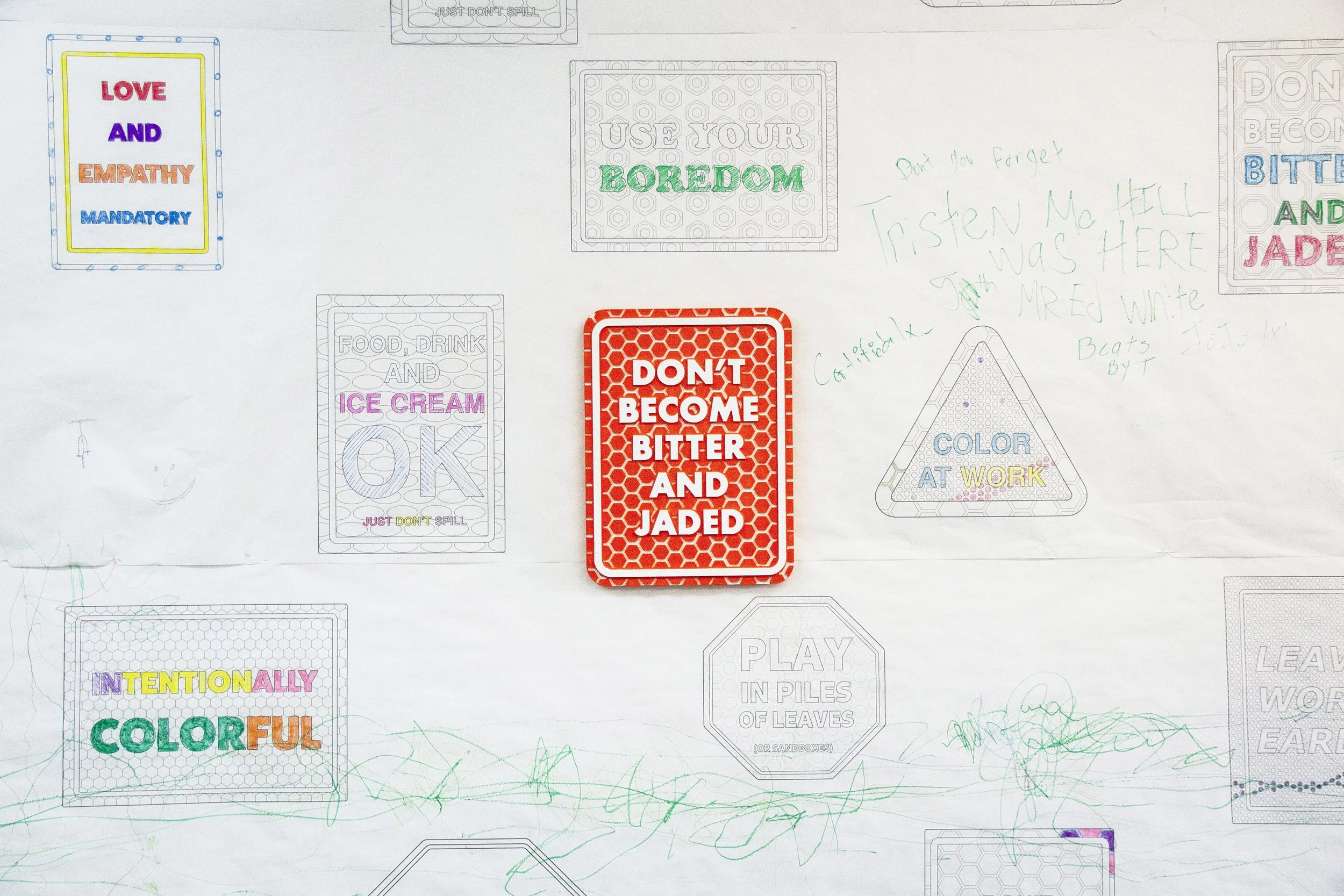 An installation image of an imitation street sign that says "Don't Become Bitter and Jaded" in white with orange hexagons in orange around the text. The artwork is installed on a wall that has white outlines of similar signs that have been colored in