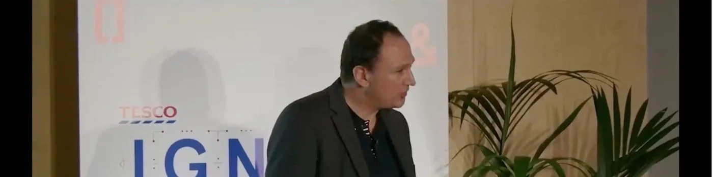 A man in a dark blazer standing next to a large polling station sign with the Tesco logo in the background.