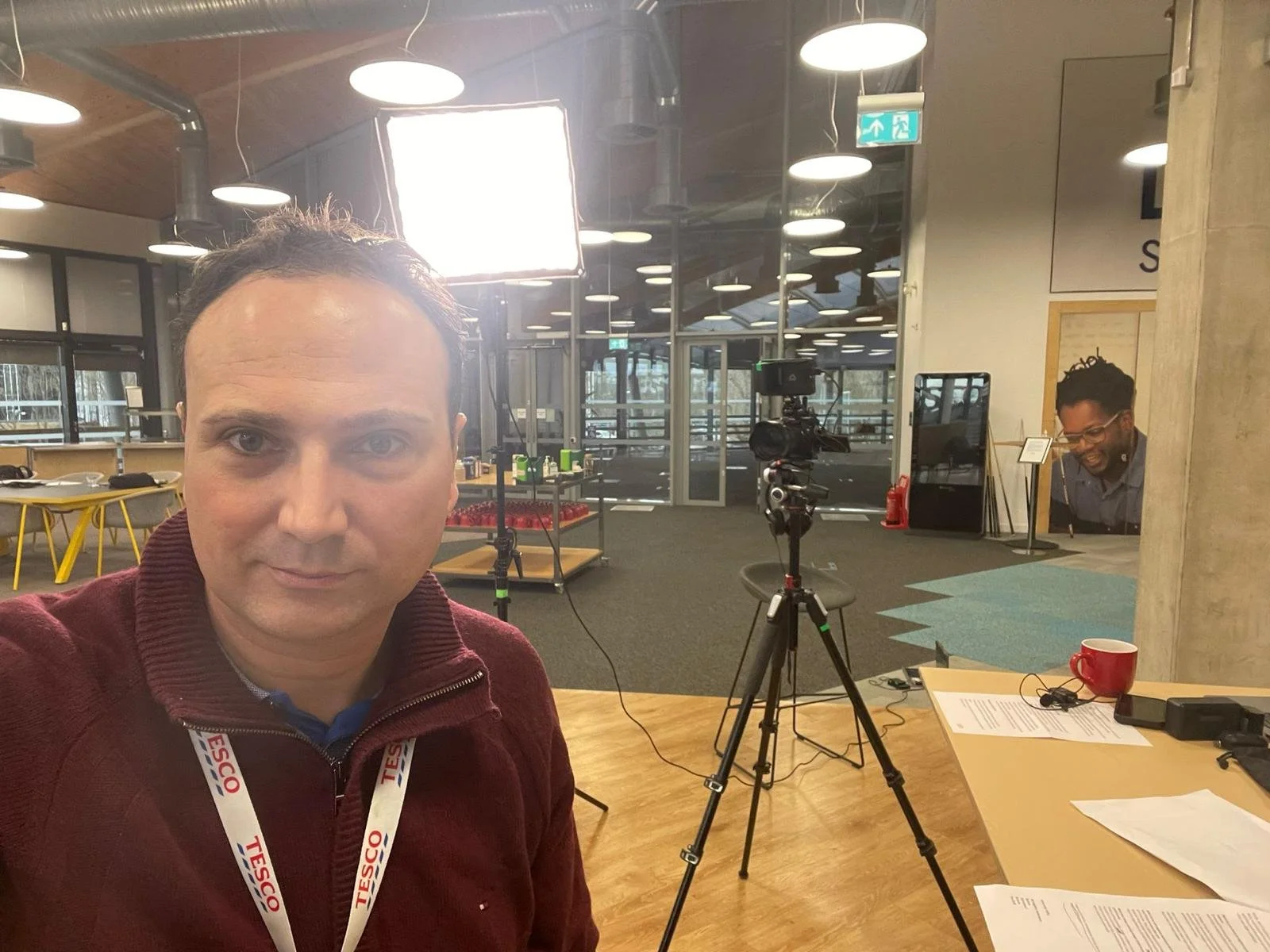 A man taking a selfie in a modern, brightly-lit indoor space with camera equipment and a colleague in the background.