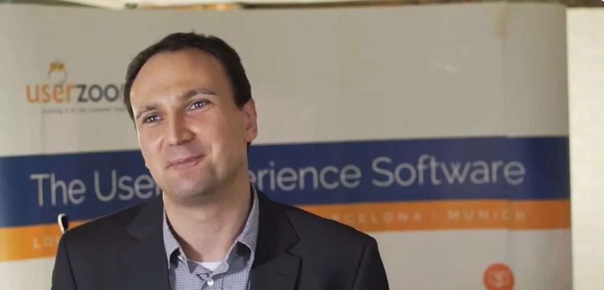 A man with dark hair and a slight smile, wearing a suit jacket over a collared shirt, standing in front of a banner that reads 'The User Experience Software' and features the logos of 'user zoom' and other text.