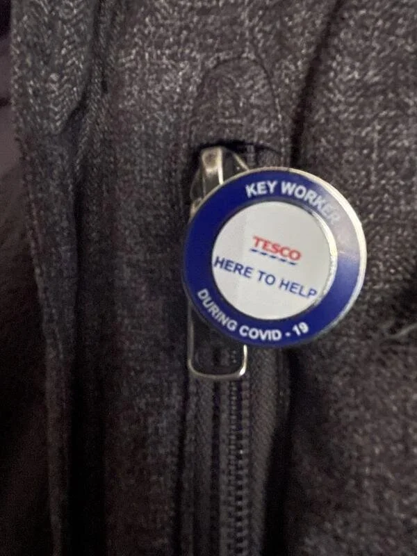 Close-up of a Tesco key worker badge attached to a dark fabric jacket, with the badge reading 'HERE TO HELP DURING COVID-19'.
