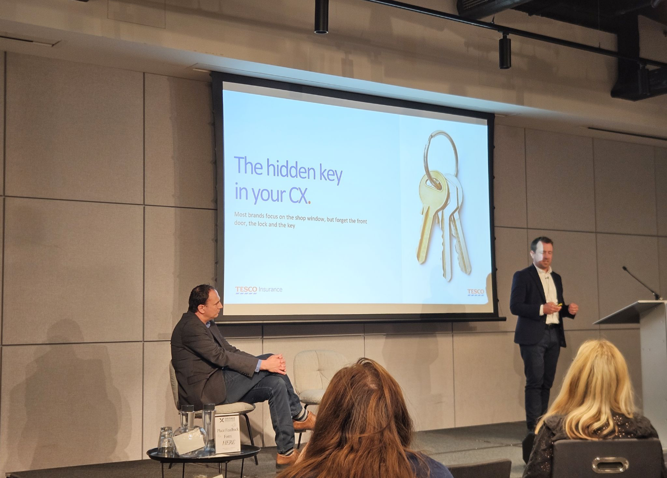 A presentation slide shows the title 'The hidden key in your CX.' and an image of three keys on a keyring. A speaker stands to the right of the screen, and another person is seated on the left side of the stage. Audience members are visible in the foreground.