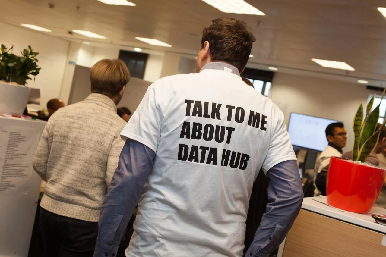 Person wearing a white T-shirt with bold black text that reads 'TALK TO ME ABOUT DATA HUB' at an indoor event or conference with other attendees and office decor.