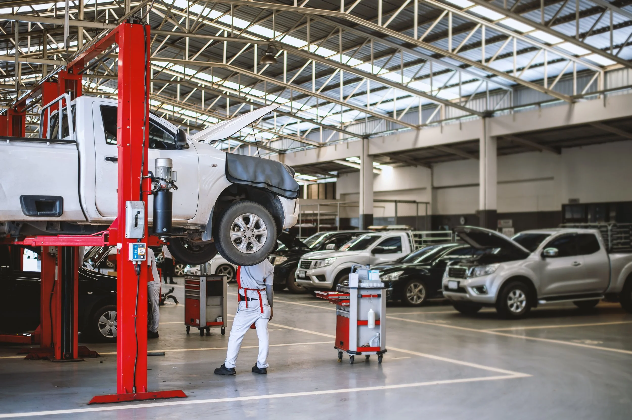 Automotive repair shop with a car on a hydraulic lift and mechanics working underneath, surrounded by other parked vehicles.