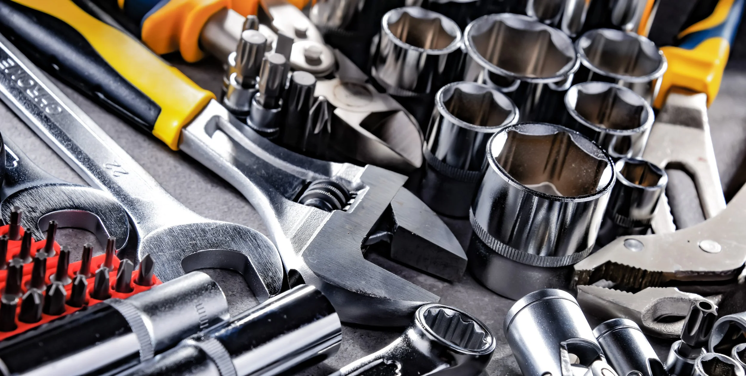 A collection of various metal tools including socket wrenches, ratchets, and screwdriver bits. Some tools have yellow and black handles, and the tools are laid out on a work surface.