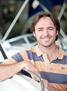 A young man with brown hair and a beard, wearing a striped polo shirt, smiling and leaning on a boat in a marina