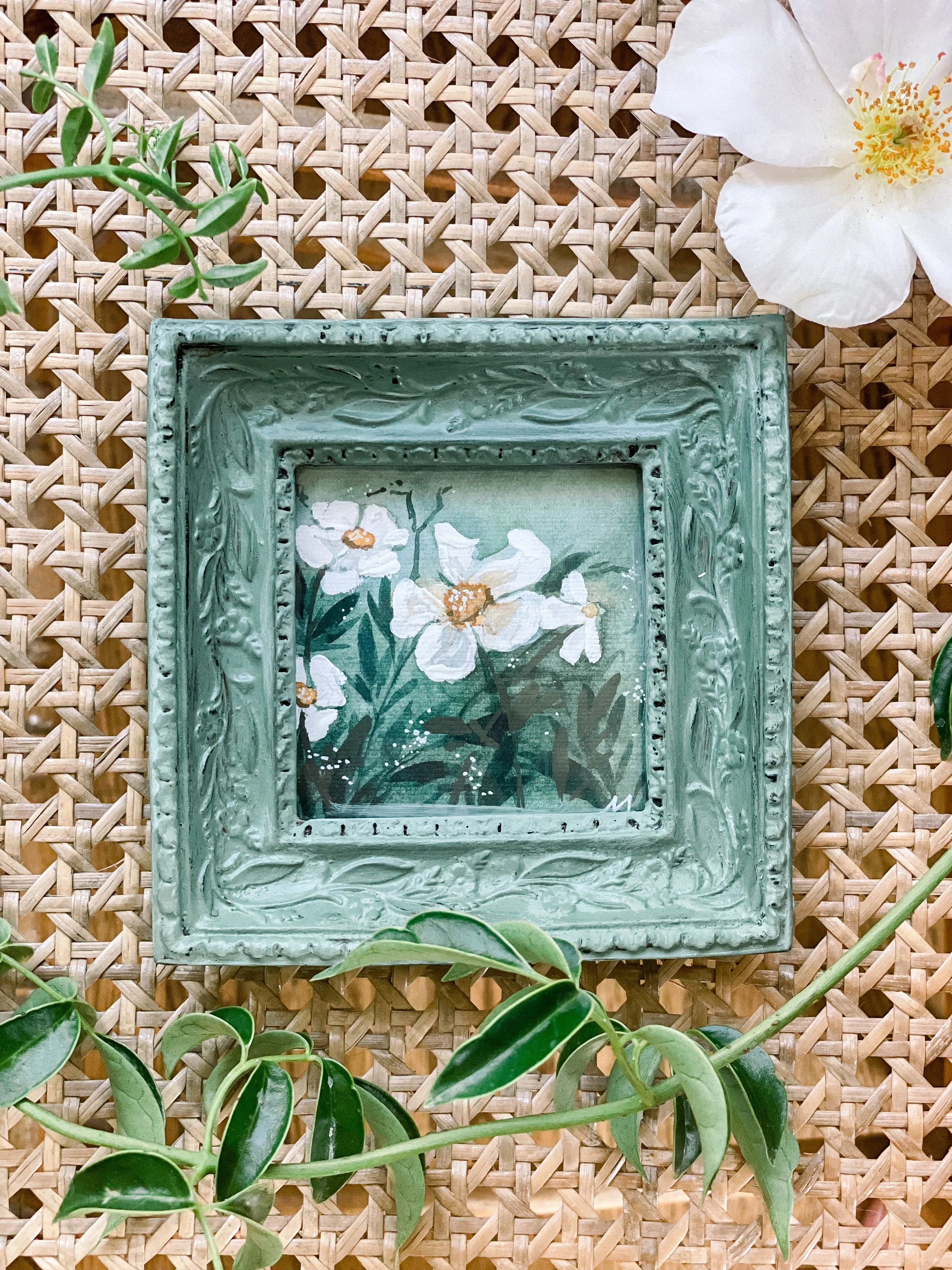 A decorative green frame with a floral painting inside, placed on a woven rattan surface. Surrounding the frame are green leaves and a white flower with yellow accents.
