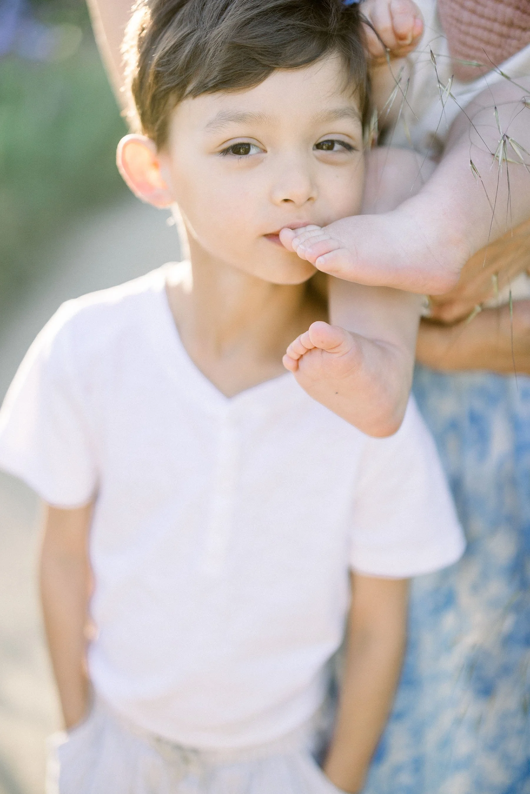 A young boy wearing a white t-shirt with his left hand behind his back, standing outdoors with a blurred background. An adult's hand is holding the boy's foot near his face, with the boy gently touching his lips with his finger, looking directly at the camera.