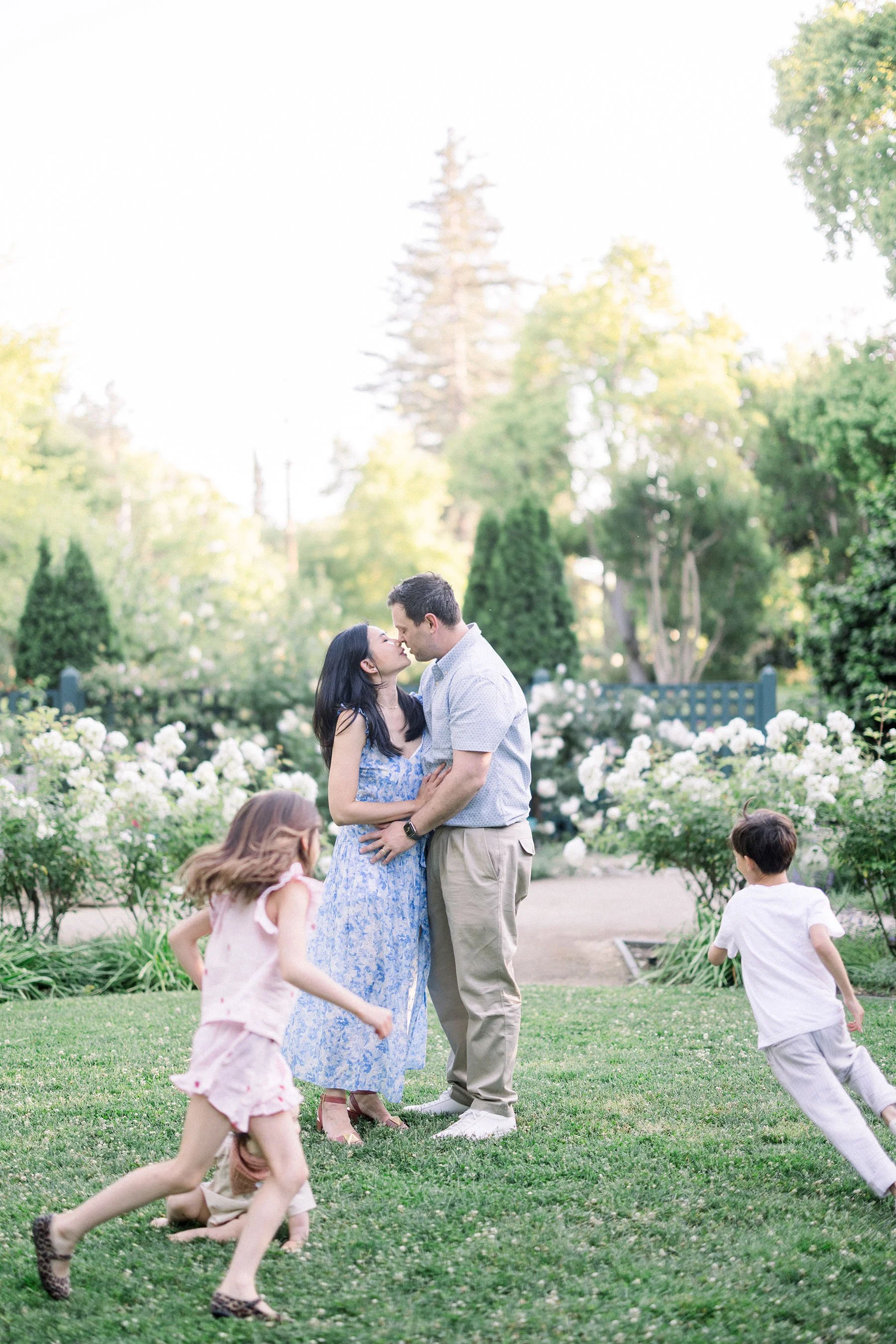 A couple stands close together, about to kiss, in a garden with white flowers and green trees, while three children run and play nearby.