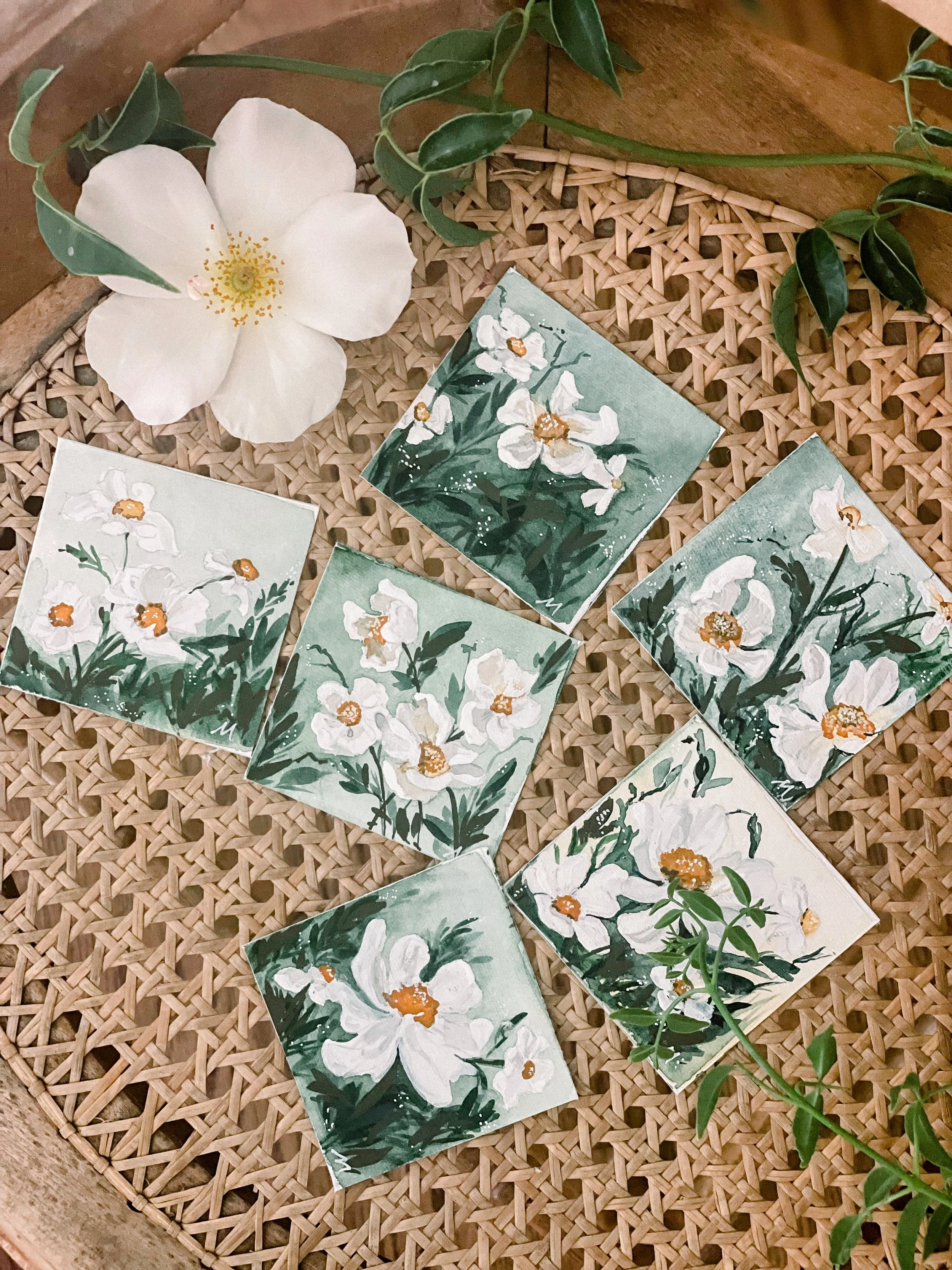 Six small painted tiles with white flowers and green leaves on a woven surface, surrounded by green leaves and a large white flower.
