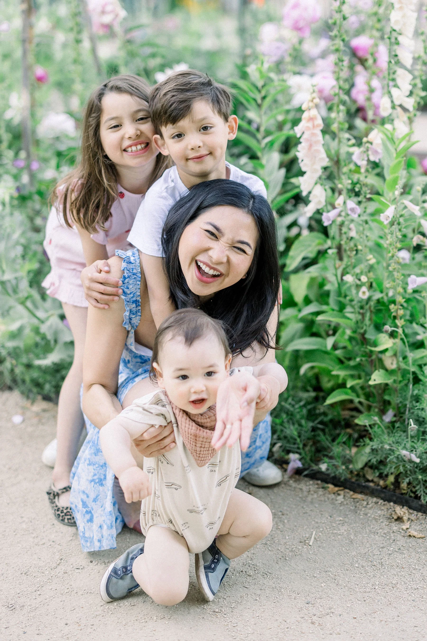 Five children, of varying ages and ethnicities, are smiling and playing together in a flower garden. They are stacked in a human pyramid with the youngest at the bottom and the older children at the top, all enjoying a sunny day outdoors.