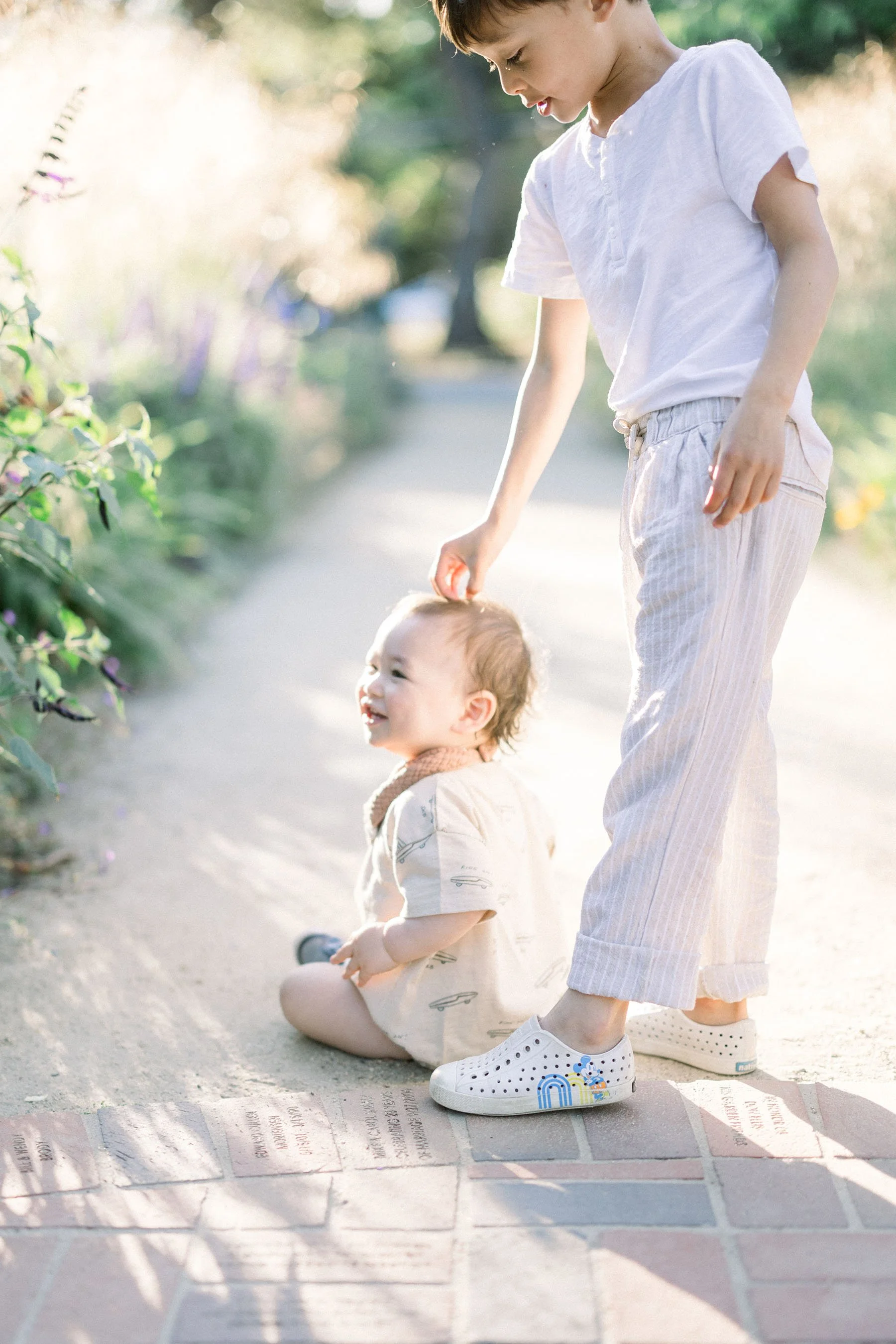A young woman and a toddler sitting on a sidewalk, with the woman gently touching the child's head. The scene is outdoors during daylight, with greenery and flowers in the background.