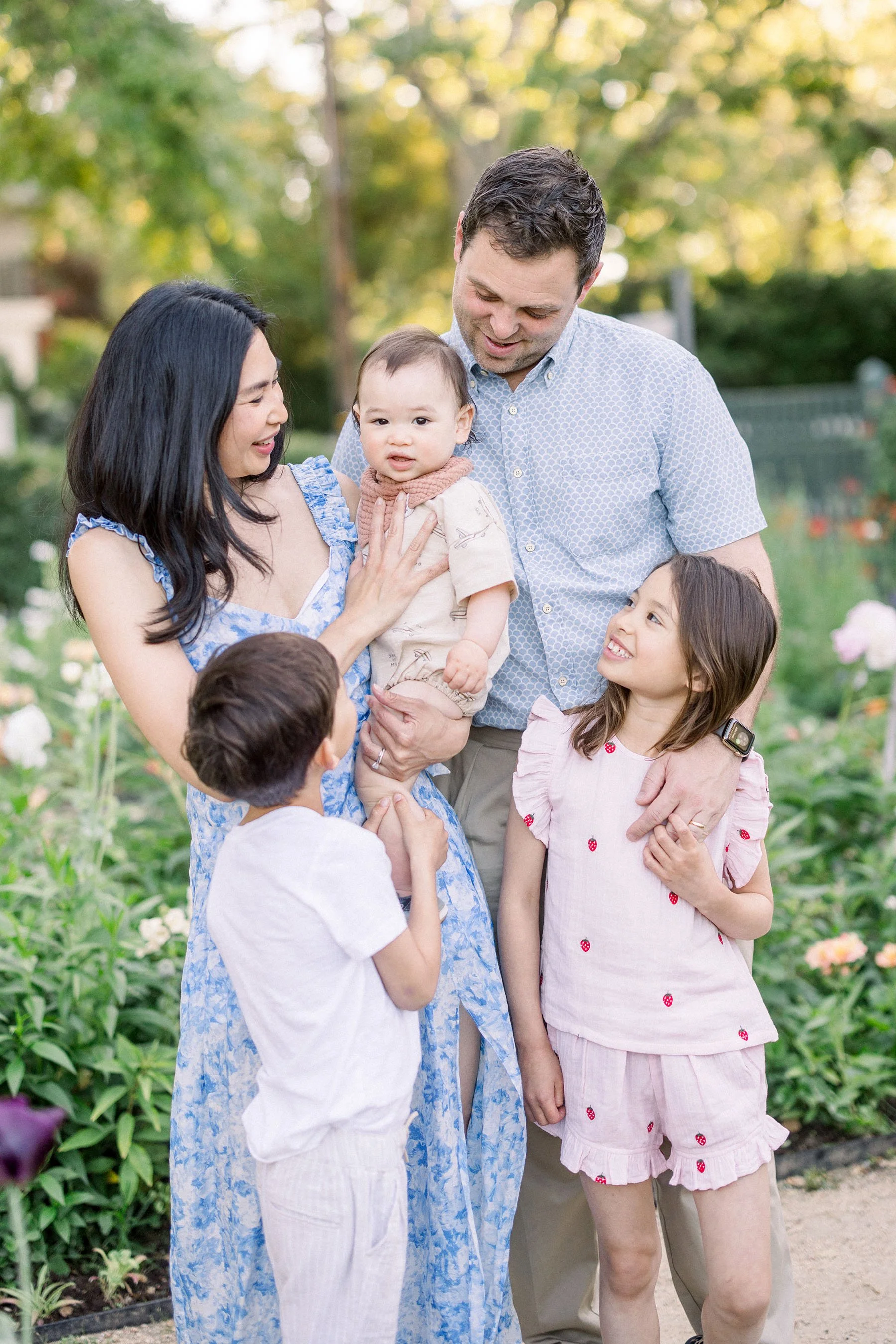 A family of five, including two parents and three children, standing in a garden with green plants and flowers, smiling and enjoying each other's company.
