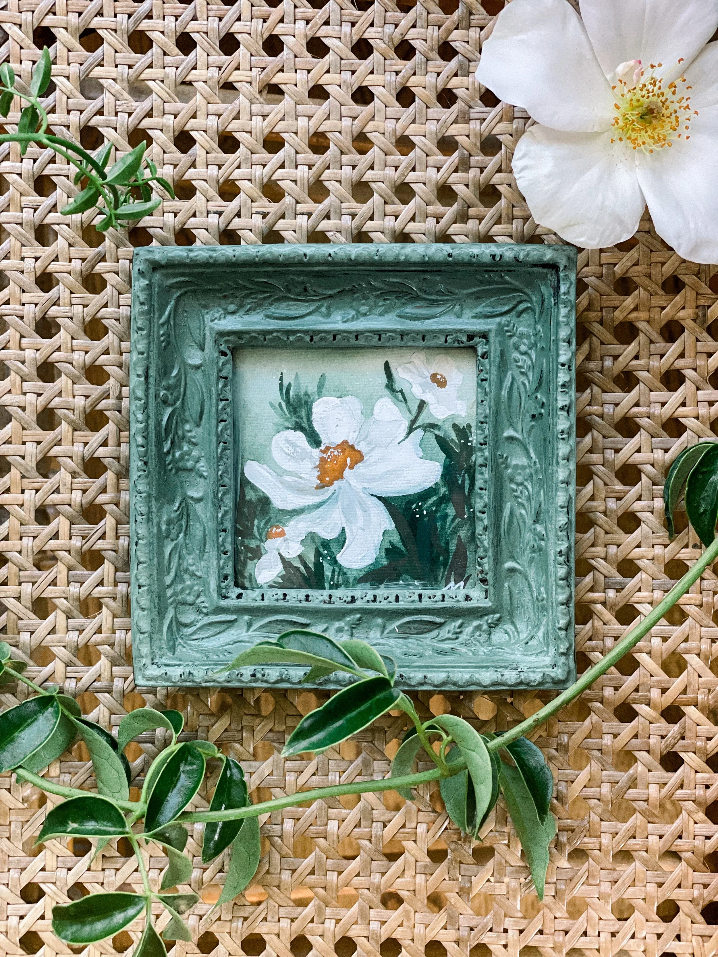 Decorative green ornate picture frame with floral painting inside, surrounded by white and yellow flowers and green leaves on a woven surface.