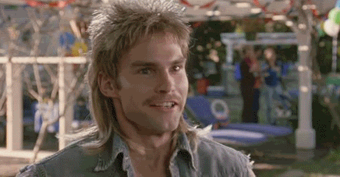 A man with long, tousled blonde hair and sideburns smiling, standing outdoors with blurred background of a park or fair.